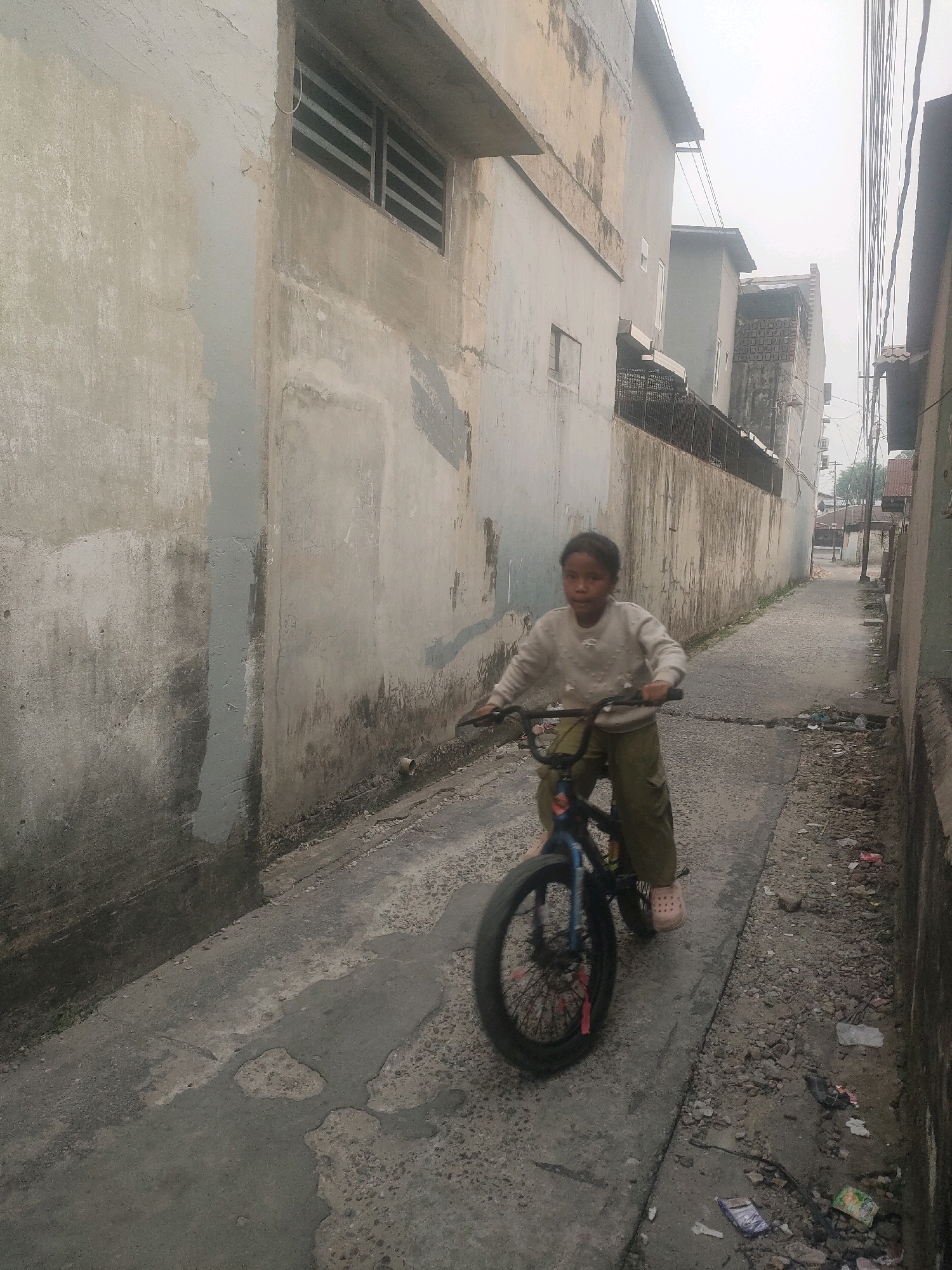 A young girl, appearing to be of Southeast Asian descent, rides a BMX bicycle down a narrow, unpaved alleyway. She is wearing a light-colored long-sleeved shirt and olive-green pants, with light pink Crocs on her feet. The bicycle has a blue frame and black wheels, with a red decoration visible on the front wheel. The alleyway is flanked by textured, aged concrete buildings, one with a window featuring horizontal slats. The ground of the alley is a mixture of concrete and dirt, with scattered debris and small puddles. In the background, more buildings and some vegetation are visible under an overcast sky. The overall impression is of a residential area in a city, potentially Pekanbaru, Indonesia, characterized by its weathered infrastructure and utilitarian construction.