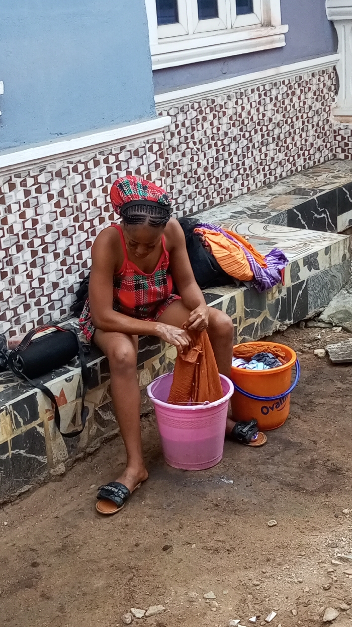 In Agburu Uke, Nigeria, a young woman is depicted hand-washing clothes outdoors during the daytime. She is seated on a low, tiled ledge, with her head bent in concentration over a task. She wears a vibrant, patterned headwrap over her braided hair, a red and green plaid camisole top, and red shorts. Black slides, one with "Supreme" visible, are on her feet.
In front of her is a pink bucket filled with water, where she is actively wringing or scrubbing a piece of orange/brown fabric. To her right, an orange bucket contains more clothes, with the brand name "Ovaltine" clearly visible on its side. Another pile of folded clothes, including orange and purple garments, rests on the tiled ledge behind the orange bucket. A black bag with a strap is placed on the ledge to her left.
The setting is against a building featuring a blue-painted upper wall and a lower section adorned with intricate, geometric brown and white tiles. A white window frame is visible on the blue wall. The ground beneath her is bare earth, suggesting a common outdoor area. The lighting indicates a clear day. The woman appears focused and diligent in her activity.