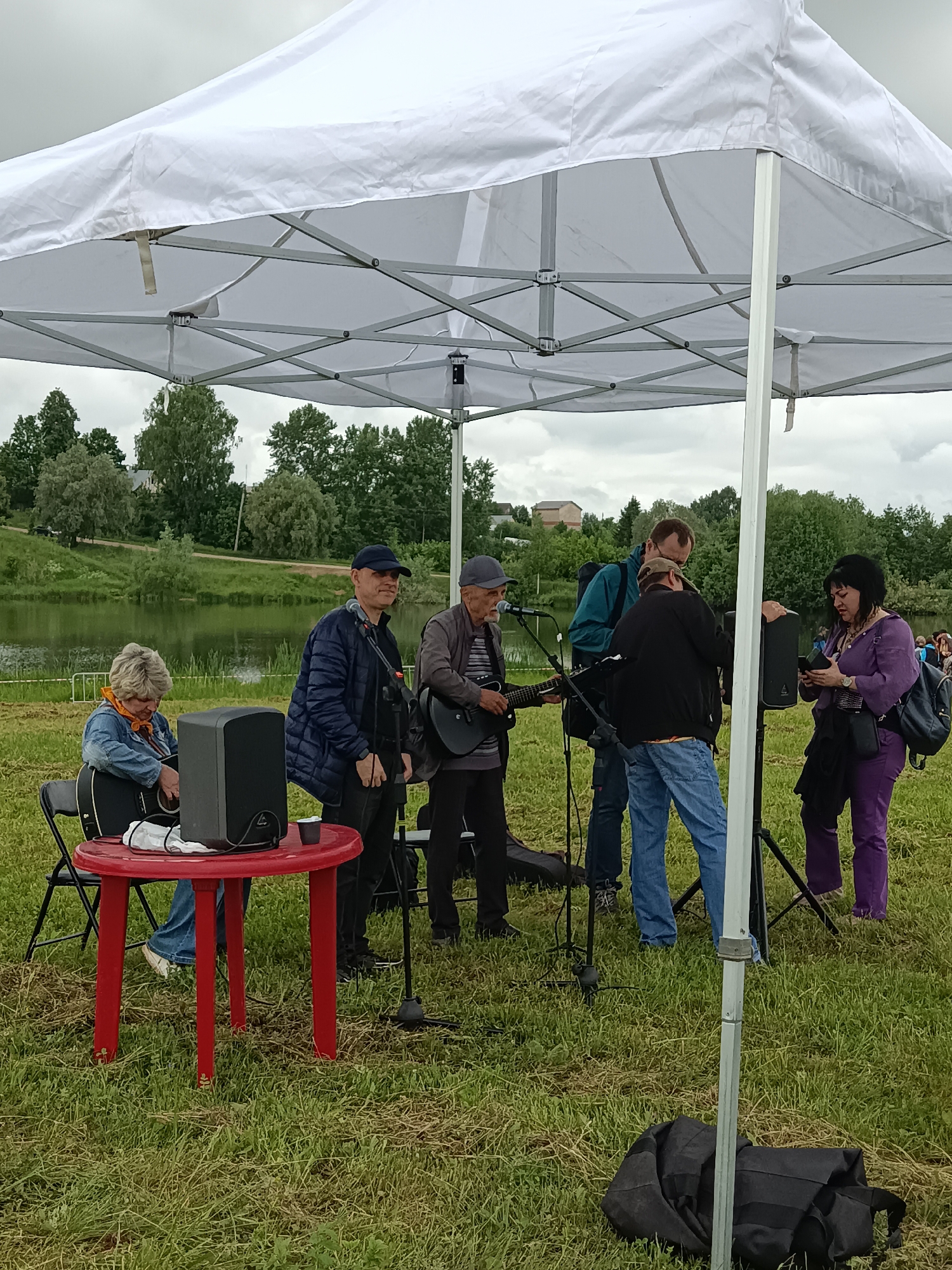 The image depicts an outdoor gathering or musical performance under a white pop-up canopy tent on a cloudy day. The setting is a grassy field beside a calm body of water, possibly a lake or river, with a tree-lined far bank and distant buildings.
Under the tent, a group of six individuals is present with musical and sound equipment. From left to right:
1. A woman with short, blonde hair and an orange scarf, wearing a denim jacket and blue jeans, sits on a black folding chair. She holds an acoustic guitar on her lap and appears to be adjusting it. A black speaker rests on a small, red, round plastic table in front of her. A white cloth and a dark cup are also on the table.
2. A man wearing a navy blue baseball cap and a dark blue puffer jacket stands, facing slightly right, with a microphone on a stand in front of him.
3. A man with a grey baseball cap and a dark jacket over a striped shirt stands to the right of the previous man, holding and playing a dark-colored electric or bass guitar, looking down at the instrument.
4. Another man, wearing a teal-colored jacket and a light-colored cap, stands with his back largely to the camera, facing toward sound equipment.
5. A man in a black jacket and blue jeans stands, adjusting equipment near a speaker stand.
6. A woman with dark hair, dressed in a purple top and trousers, stands on the far right, looking down at a small black electronic device, likely a smartphone, in her hands. She has a black shoulder bag and a smaller cross-body bag.
Several black microphone and speaker stands are visible throughout the central area. The ground is covered in somewhat sparse and uneven green grass. A collapsed black bag, possibly a carrying case for equipment, lies on the grass near the right-most tent support pole. The sky above the tent is overcast with light grey clouds. A narrow path or road is discernible on the far bank of the water, traversing between the waterline and the trees.