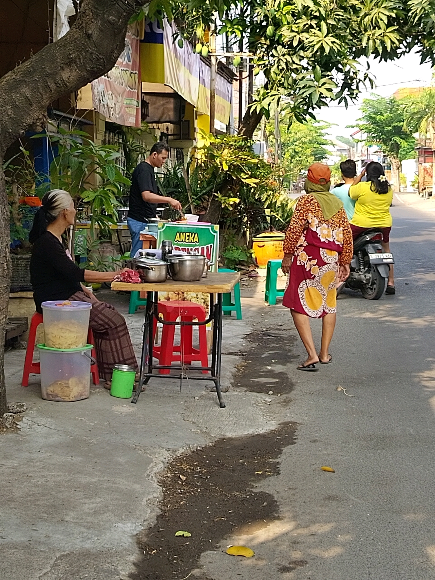 The image captures a street scene in Mojokerto, Indonesia, likely during the day, indicated by the bright sunlight and shadows. In the foreground, a woman with grey-streaked black hair is seated on a red stool, facing left. She wears a black long-sleeved shirt and a patterned skirt. In front of her, on a wooden table, are two metal pots, a pile of red cloth, and a green plastic container. To the left of the table, stacked plastic containers hold what appears to be food items, possibly noodles. A man in a black t-shirt and blue jeans stands behind the table, seemingly engaged in preparing food, with a sign that reads "ANEKA" in green.
Further back, three individuals are walking away from the camera. The first, wearing a patterned dress and an orange headscarf, is in the center. Behind her, a person in a light blue t-shirt is visible. The furthest person, wearing a yellow t-shirt, is on the right, next to a parked motorcycle. The background reveals shops and buildings along the street, with lush green trees providing shade. The road surface is asphalt, showing some wear and patches of water. The overall impression is of a bustling local market or roadside food stall area.