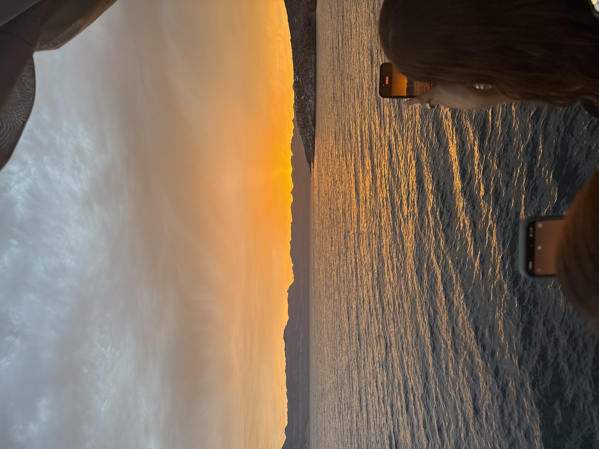 A horizontal image captures a scene during sunset with a body of water, possibly an ocean or a large lake, occupying the right half. The water reflects the warm hues of the setting sun, creating a shimmering effect. The left half of the image features the sky transitioning from soft, warm colors near the horizon to cooler tones higher up. A silhouette of a landmass, potentially a mountain range or coastline, separates the sky from the water.
Two people are visible, each holding a smartphone. The first person on the upper right side is actively filming or taking a photo, as indicated by the recording icon on their phone screen. The second person is located on the lower right, also holding a smartphone.
A sheer curtain hangs in the upper-left corner of the frame, suggesting the photograph was taken from inside a building or structure.
The location is Unknown, Mexico.