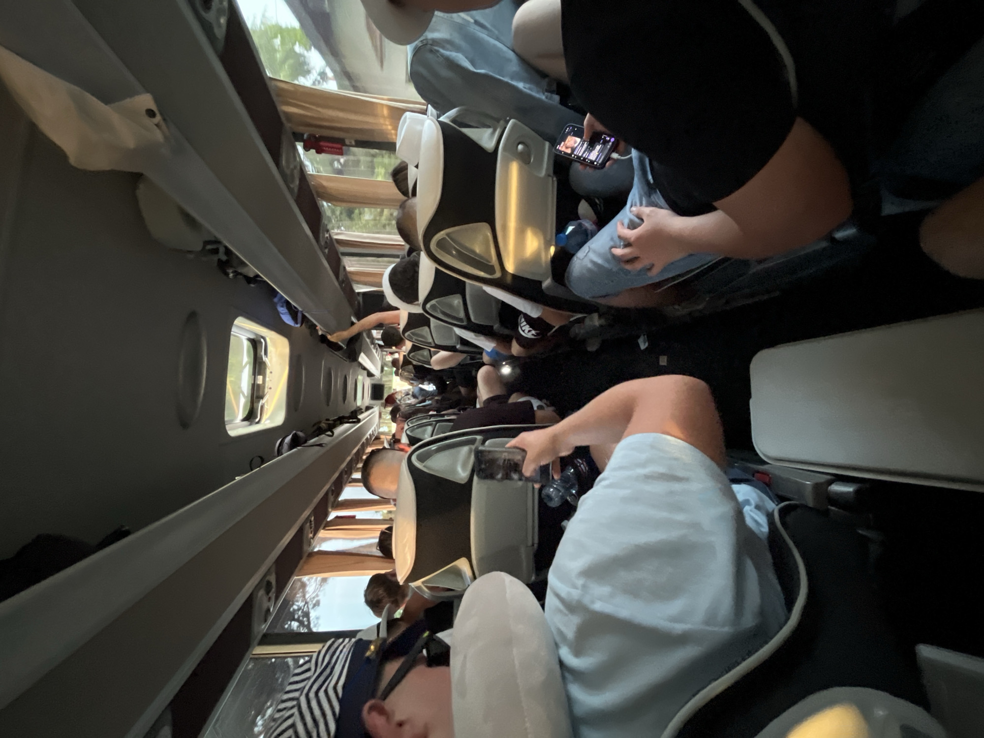 The image shows the interior of a bus, likely on a trip. Passengers are seated. Some are using mobile phones. Luggage is placed overhead. The location of Ca' Savio, Italy is not verifiable from the image.