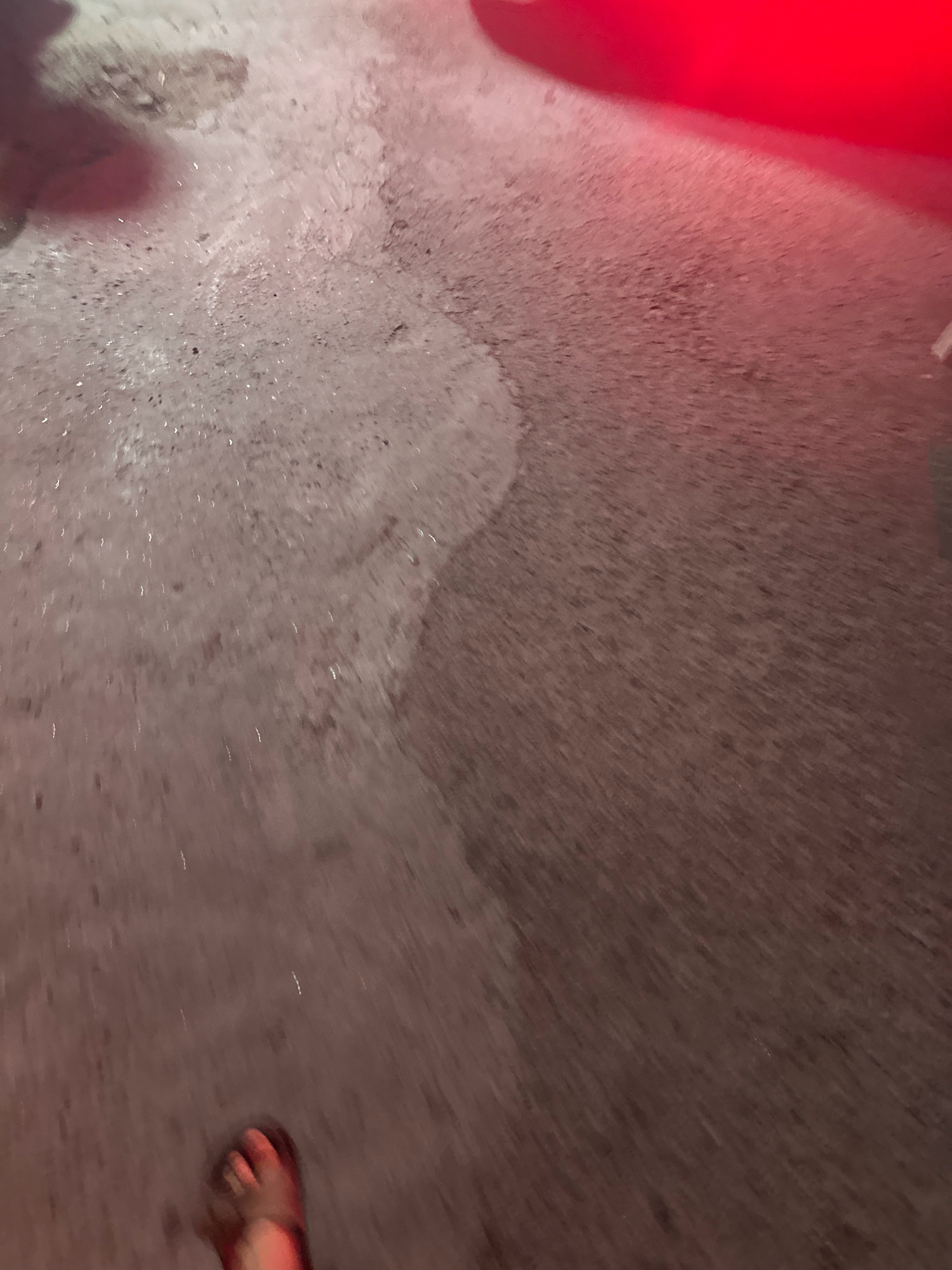 The image depicts a close-up, ground-level view of a paved surface, possibly a street or sidewalk, with a person's right foot, wearing a sandal, visible at the bottom. The ground appears wet in some areas, creating a sheen, and there are some scattered debris or small stones. A bright red light source, likely from a vehicle or signage, casts a strong red hue across the upper right portion of the scene and illuminates the wet ground. The overall impression is that of an outdoor urban environment, possibly at night or in low light conditions. The location is identified as Lalitpur, Nepal.