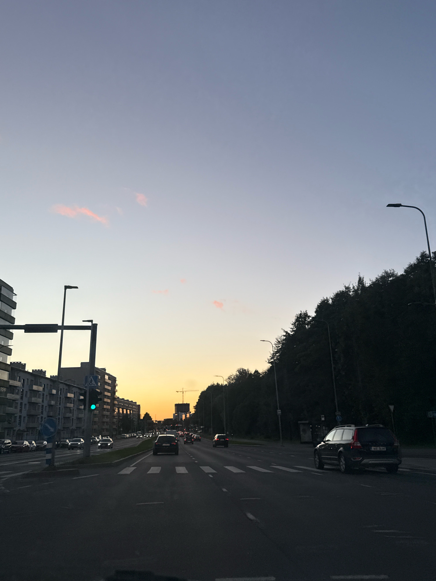 The image captures a street scene in Tallinn, Estonia, during twilight. The sky is a gradient of pale blue and soft orange, with a few wisps of pink clouds. On the left side of the road, modern apartment buildings line the street. Traffic lights, with a green light illuminated, are visible. Several cars are driving on the multi-lane road. On the right side, a dense line of trees creates a dark silhouette against the sky. Streetlights are sparsely placed along the road. In the distance, a construction crane stands against the twilight sky. The overall atmosphere is that of an urban environment transitioning from day to night.
