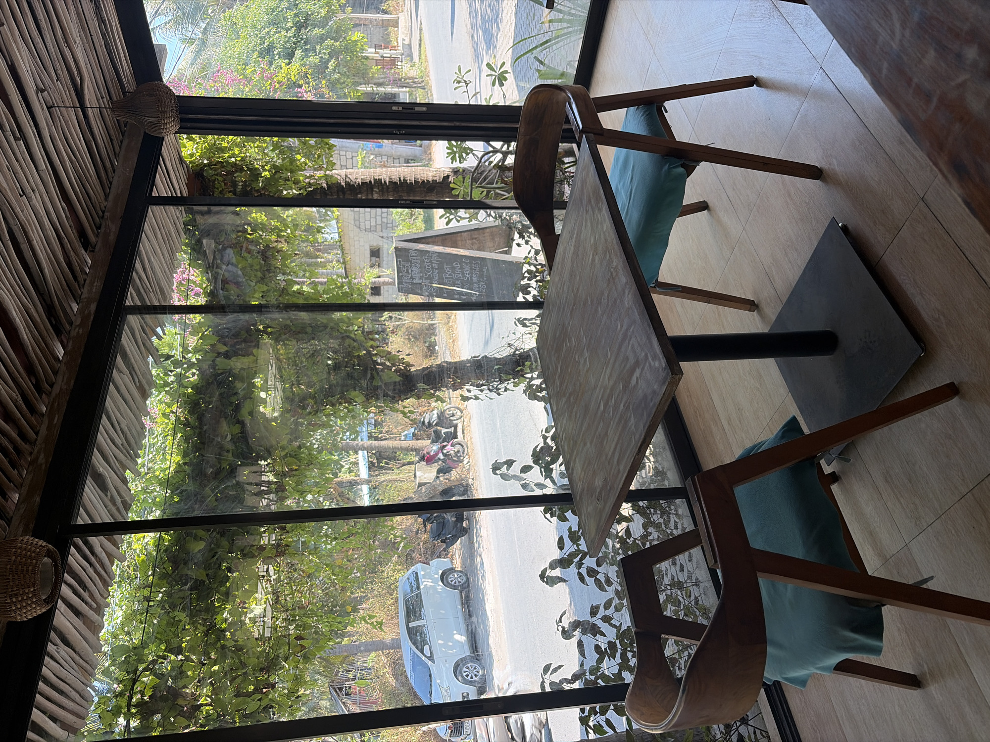 The image shows a cafe interior with large windows overlooking a street scene in Nusa Penida, Indonesia. Inside, there is a square wooden table with two wooden chairs, each with a light blue cushion. One chair is positioned to the left of the table, and the other to the right. The table has a black metal pedestal base. Beyond the table and chairs, the large windows offer a view of the exterior.
Outside the cafe, a white minivan is parked on the side of a paved road. Behind the minivan, two motorcycles are parked. Lush green foliage hangs over the window, partially obscuring the view of the street and the surrounding vegetation. A wooden A-frame sign with chalk writing is visible on the right side of the exterior view. The ceiling of the cafe is constructed of wooden beams, and a woven basket-style light fixture hangs from the ceiling. The floor inside the cafe appears to be tiled with light brown square tiles. The overall impression is a relaxed, open-air dining or seating area with a view of the local environment.