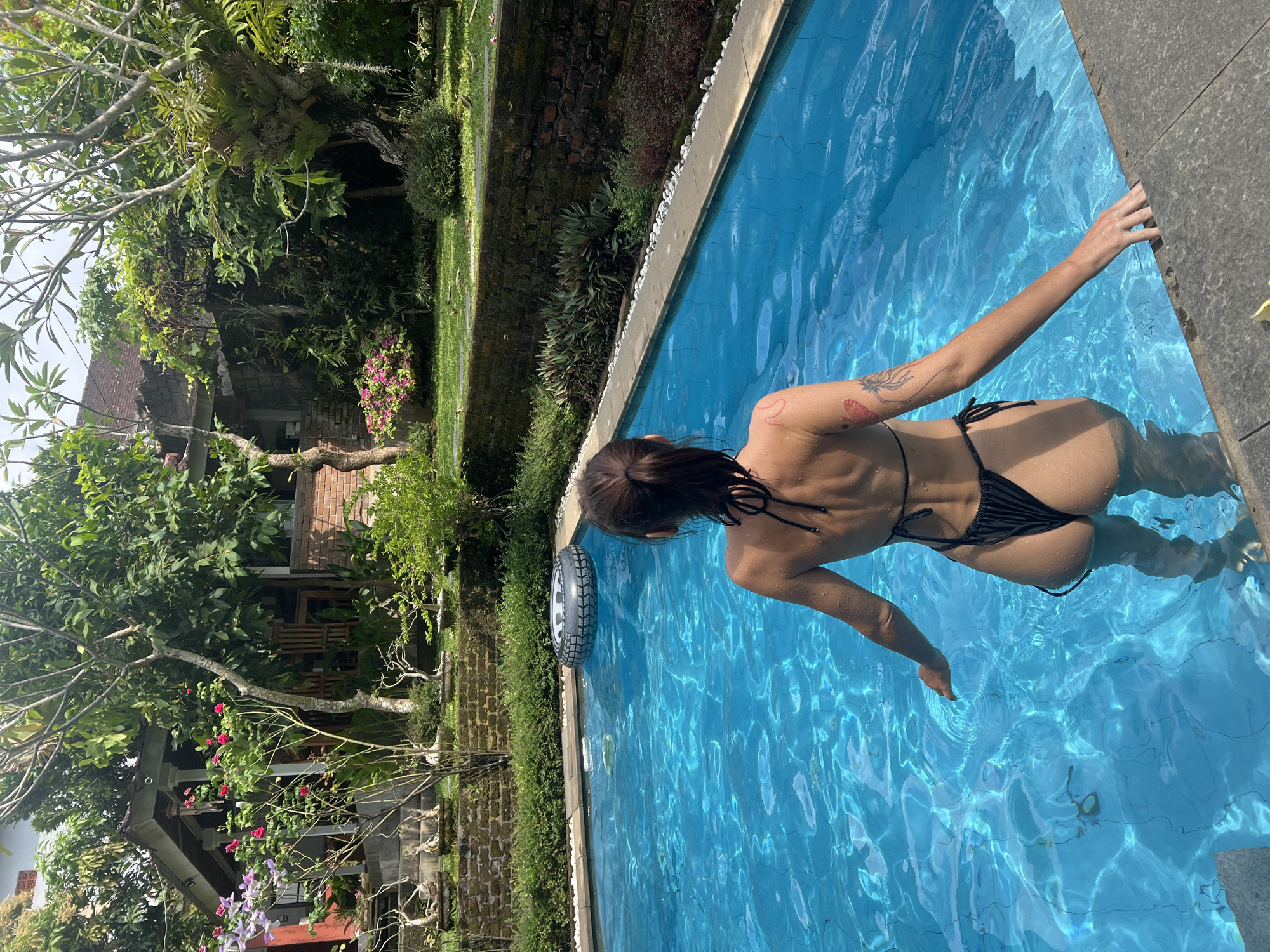 A woman in a black bikini is in a blue swimming pool. She has short brown hair and a tattoo on her upper back. The pool is bordered by stone tiles and white stones. A decorative car tire is next to the pool. Behind the pool is a stone wall covered in greenery and a building with trees and flowers. The image was taken in North Kuta, Indonesia.