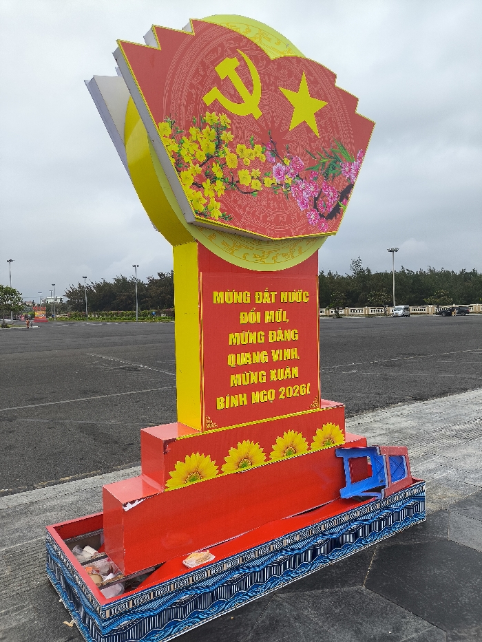The image displays a large, elaborate monument positioned in a paved open area under an overcast sky. The monument consists of a multi-tiered base, a central column, and a prominent fan-shaped top section.
The base features red stepped platforms adorned with yellow sunflower decals. Below this, a rectangular planter-like structure with blue and white wave patterns contains various small, indistinct objects. A small, bright blue plastic stool is partially resting on the lower base structure, leaning against the red steps.
The central column is predominantly red with yellow borders, displaying vertical text in Vietnamese: "MỪNG ĐẤT NƯỚC ĐỔI MỚI, MỪNG ĐẢNG QUANG VINH, MỪNG XUÂN BÍNH NGỌ 2026!"
The fan-shaped top section is red and yellow, featuring a large yellow hammer and sickle symbol alongside a yellow five-pointed star, indicative of Vietnamese communist iconography. Below these symbols, colorful depictions of yellow apricot blossoms and pink peach blossoms are present. This entire section is bordered by a yellow pattern.
The monument is situated on a large, empty asphalt surface resembling a plaza or parking lot. In the background, a dense line of trees, likely coastal vegetation, is visible, along with multiple tall lampposts. Several vehicles, including a white car and a dark SUV, are parked or driving in the distance. The immediate ground to the bottom right shows dark grey hexagonal paving tiles. No individuals are clearly visible in the immediate vicinity of the monument.
This scene is located in Thành phố Tuy Hòa, Vietnam.