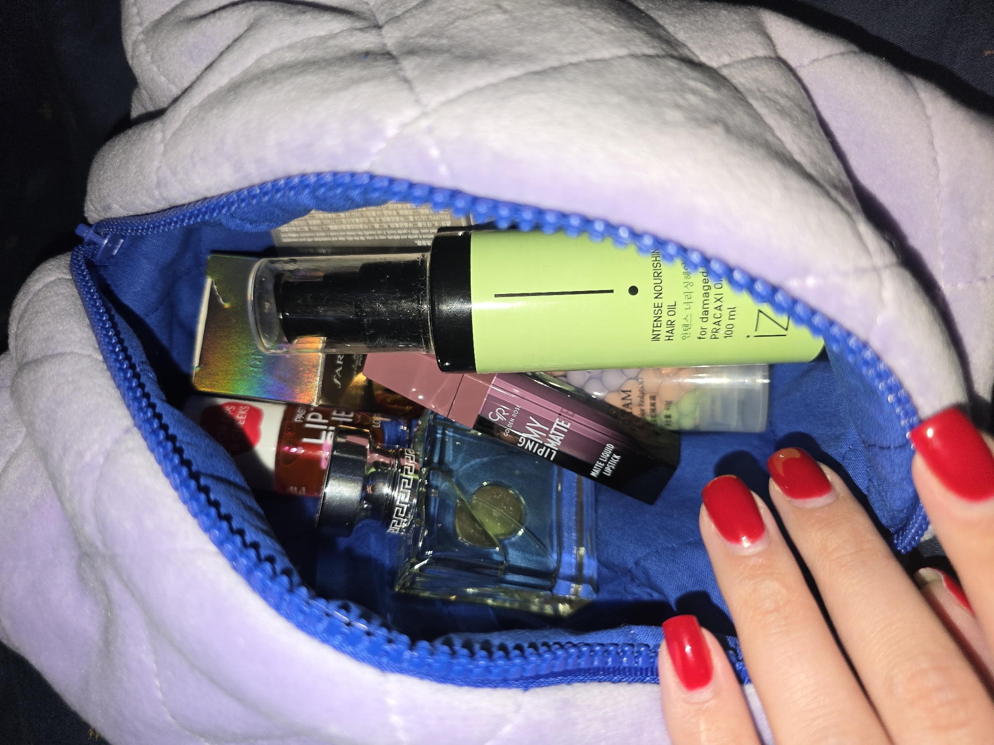 The image is a close-up, eye-level shot of a partially open lavender quilted makeup bag with a blue zipper. Inside the bag are several beauty products. In the foreground on the left, a person's hand with bright red nail polish is visible, reaching towards the bag. Prominently featured inside the bag is a glass perfume bottle with blue liquid and a silver cap, a tube of "GOLDEN ROSE LIPING MY MATTE" lipstick, and a bright green bottle labeled "INTENSE NOURISHING HAIR OIL" with Korean text. Other cosmetic items are partially visible. The background is dark and indistinct. The scene appears to be indoors, possibly in a domestic setting. There is no information in the image to suggest a specific location in Petropavlivska Borshchahivka, Ukraine.