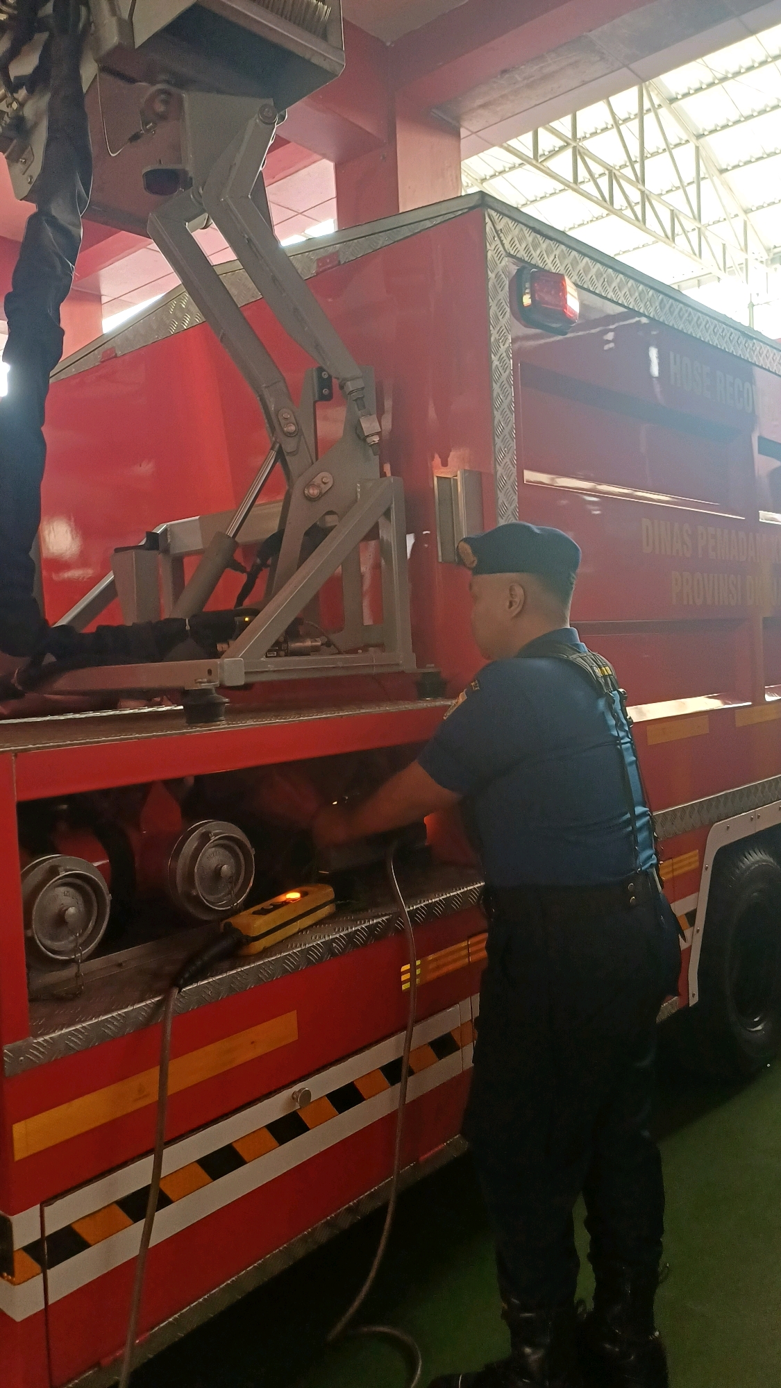 A firefighter in a blue uniform and cap is working on the side of a red fire truck. The fire truck has "DINAS PEMADAM..." and "...PROVINSI DKI" written on its side. The firefighter is using a yellow handheld device. There are two hose reels visible on the truck. The scene appears to be indoors, possibly in a fire station, with a metal roof structure visible above.