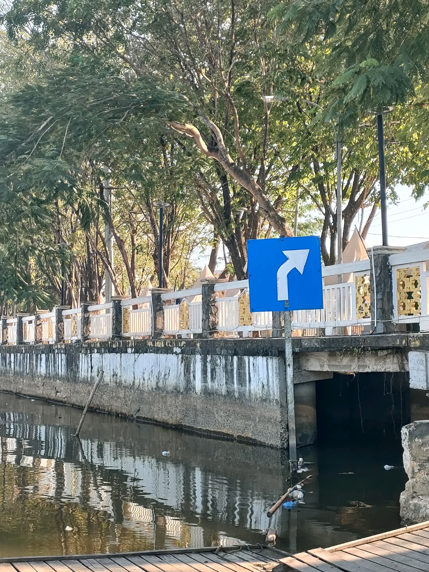 A blue square sign with a white arrow indicating a right turn is posted on a pole on the left side of the image. The sign is located in front of a white and gold railing that lines the edge of a concrete structure. Beyond the railing, a row of trees with lush green foliage is visible, with sunlight filtering through the leaves. Below the railing and concrete structure is a body of dark water, reflecting the trees and the railing. Debris, including plastic bags and bottles, floats on the water's surface, particularly near a concrete bridge in the background. The water is contained by a weathered concrete wall on the left and a concrete bridge abutment on the right. Wooden planks form a walkway in the foreground, extending from the bottom left of the frame. The scene appears to be along a canal or river within the city of Banjarmasin, Indonesia, given the characteristic waterways and urban infrastructure.