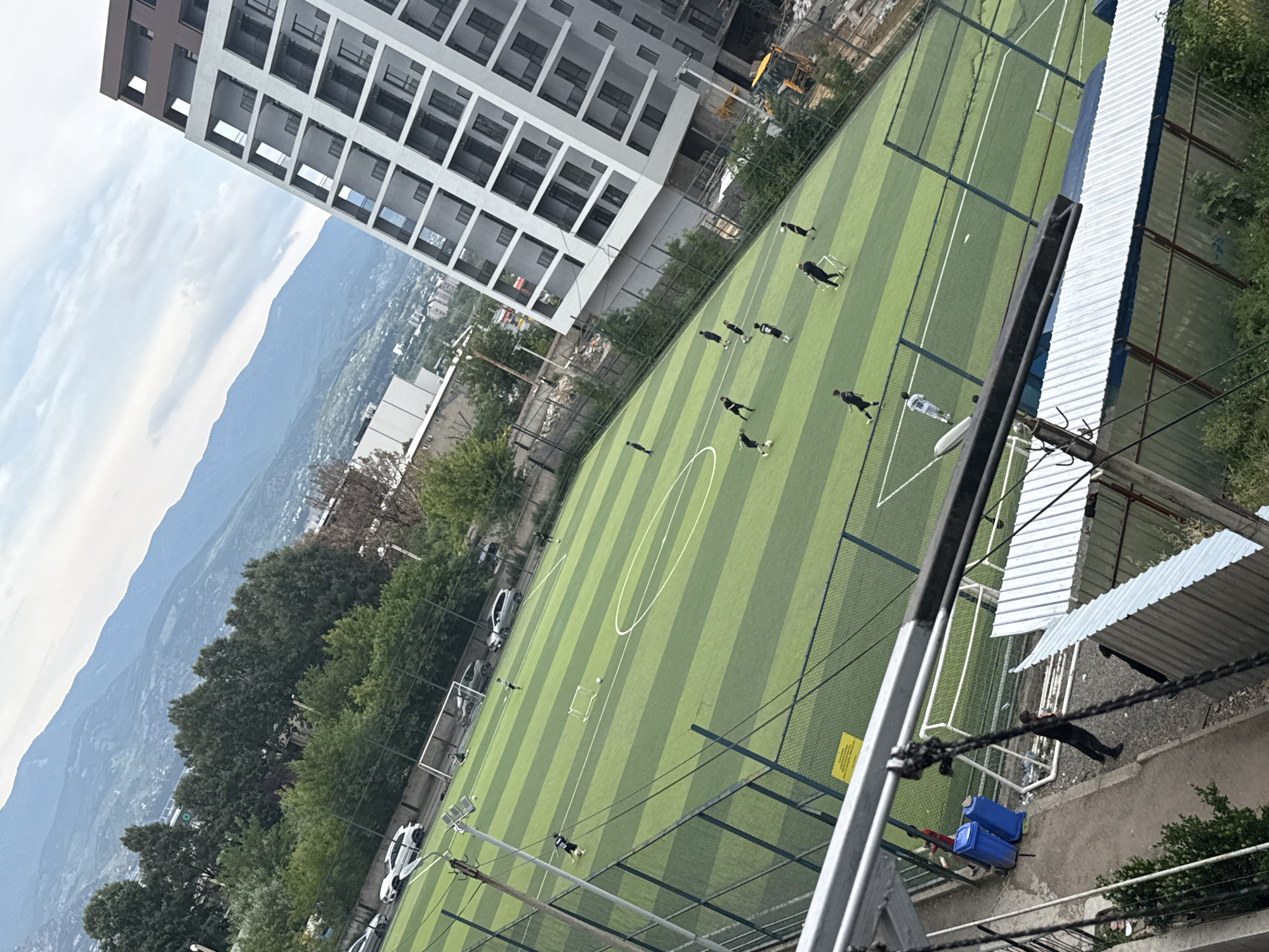This image depicts a football (soccer) field in Tbilisi, Georgia, with several individuals engaged in a game or practice. The field is artificial turf with clear markings for play. In the background, a modern, multi-story building with numerous balconies dominates the right side of the frame. Further in the distance, a range of mountains is visible under a cloudy sky, suggesting a location on the outskirts of the city or in a valley. The scene includes trees, fences surrounding the field, parked cars, and construction equipment, indicating an active urban environment. The camera angle is from an elevated position, looking down onto the field.