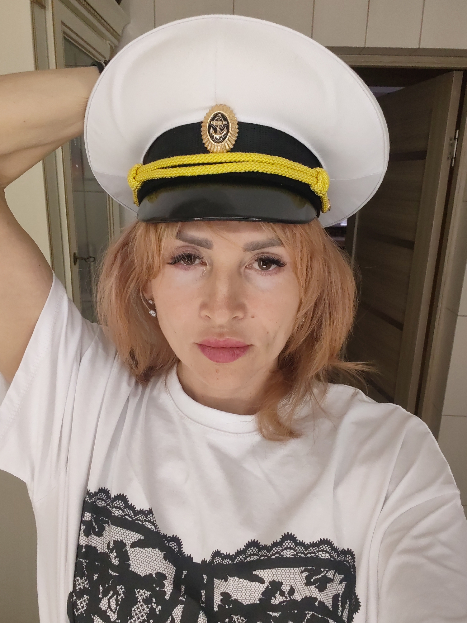 A woman with light brown hair, wearing a white t-shirt with a black lace print and a white captain's hat with a gold emblem, poses for a selfie. Her right arm is raised, holding the brim of the hat. The background appears to be an indoor setting, possibly a home, with a closed door visible on the right. The scene is well-lit, and the woman is looking directly at the camera with a neutral expression. There is no indication of the location being in Bol'shie Mkhi, Russia.