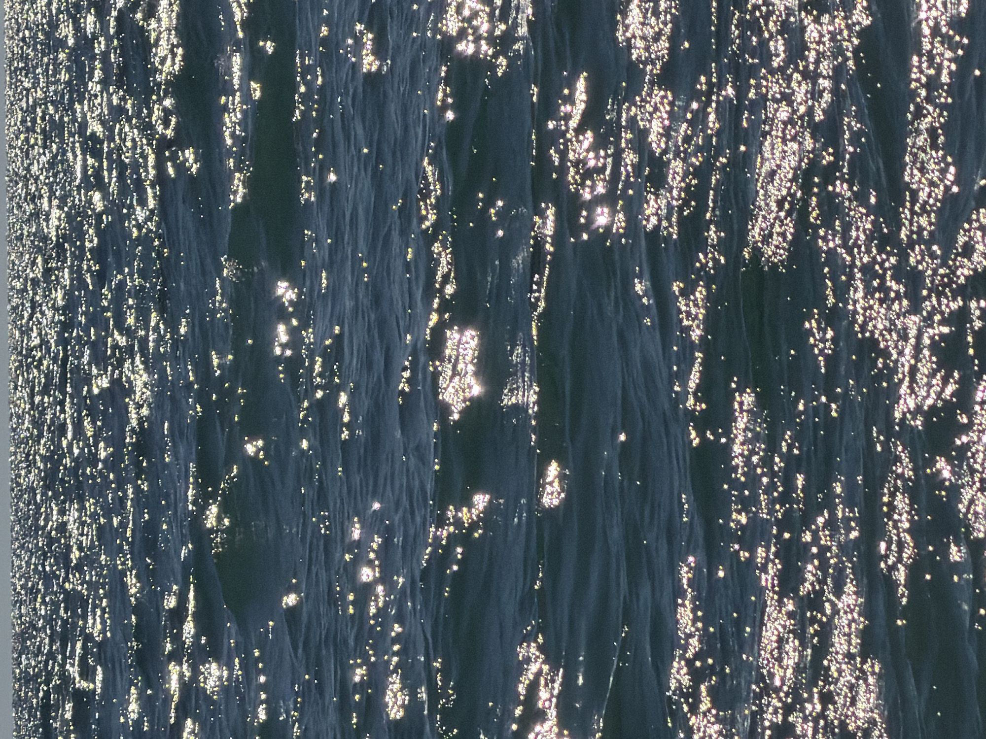 The image displays a wide expanse of dark, rippling water, likely the sea, reflecting the sun. Numerous bright, scattered glints of light are visible on the surface, indicating a sunny day and possibly a gentle breeze causing small waves. The horizon line is subtly visible in the distance, separating the water from a pale, hazy sky. There are no discernible structures, people, or specific landmarks visible that would indicate a location within the city of Sochi, Russia. The scene is solely focused on the natural environment of the water's surface.