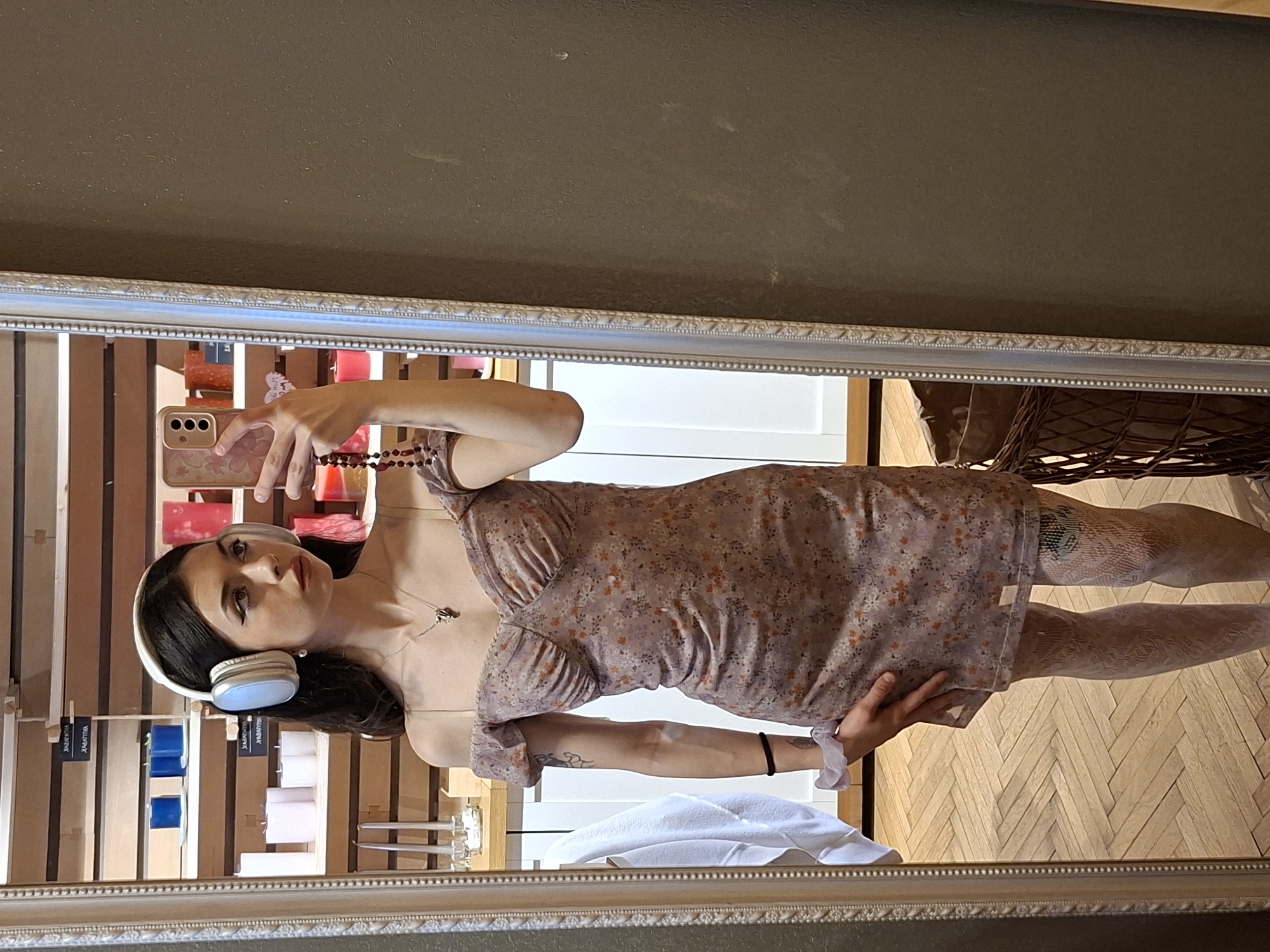 A mirror selfie shows a woman with dark hair wearing headphones, a floral dress, and patterned tights. She is holding a phone in her hand. The reflection shows a display of candles and a wooden floor. Based on the reflection, the location could be a shop or a showroom in Sofia, Bulgaria.