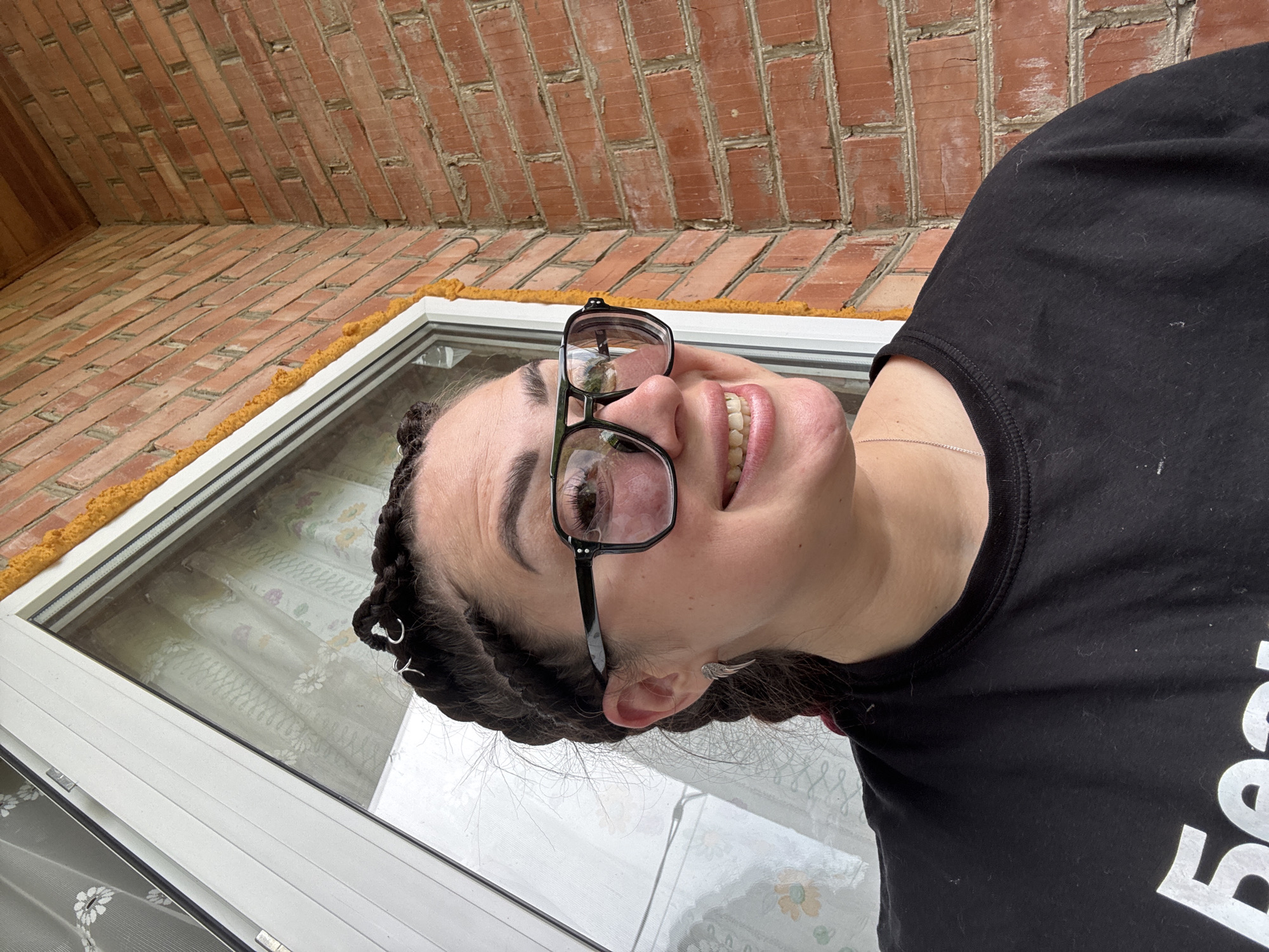 A woman with braided hair and large black-rimmed glasses smiles and looks to her right. She is wearing a black t-shirt and a silver necklace. She has a silver earring shaped like a wing in her left ear. The background consists of a brick wall and a window with sheer curtains. The scene appears to be outdoors, possibly on a porch or balcony.