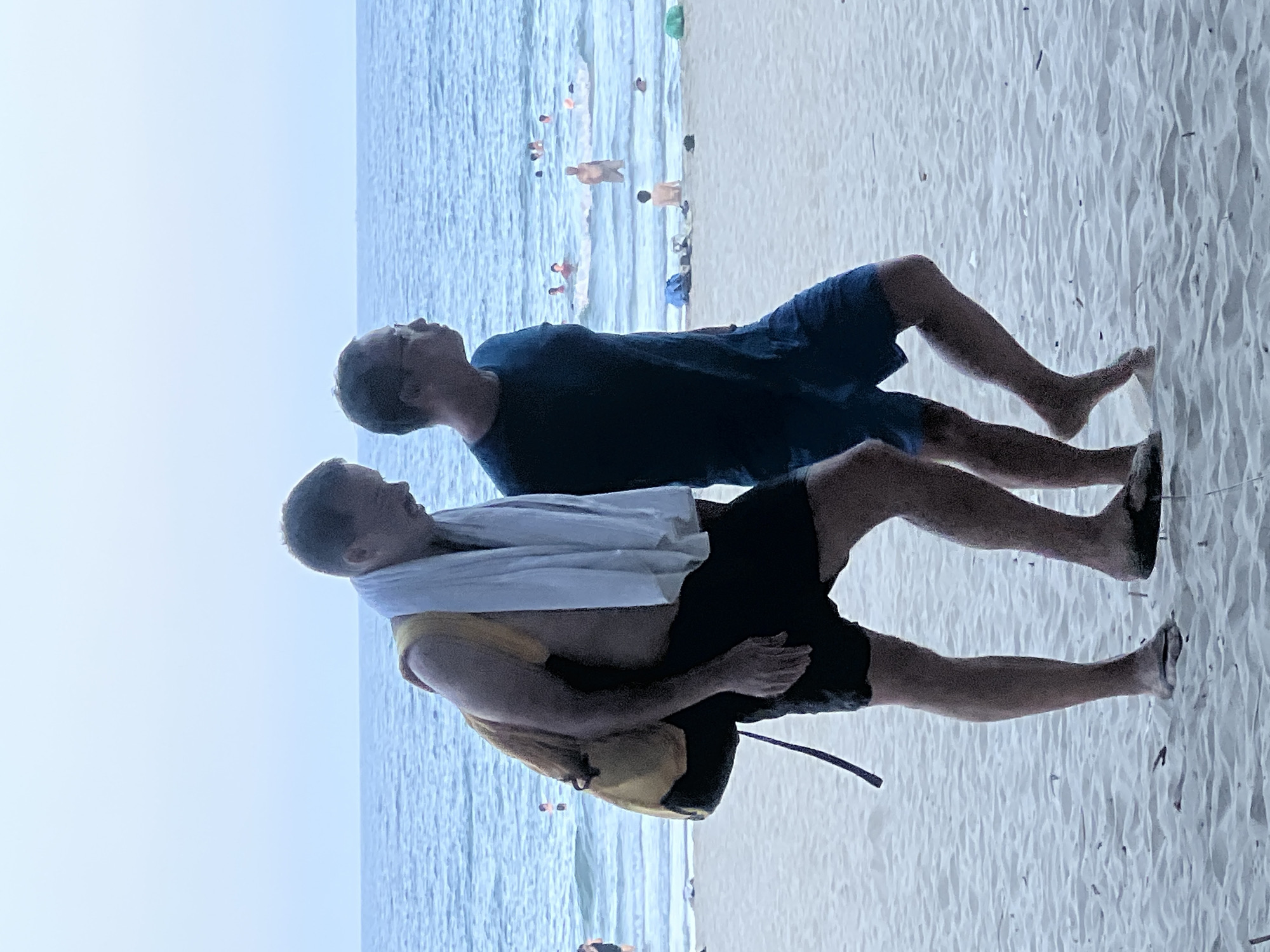 Two men walk along a sandy beach with the ocean and sky in the background. The man on the left, who is shirtless, wears black shorts and a yellow backpack slung over his right shoulder with a white towel draped over it. He is looking down. The man on the right wears a dark blue t-shirt and blue shorts. He is looking to the left. In the distance, other beachgoers are visible in the water and on the shore. The scene appears to be Đà Nẵng, Vietnam, given the presence of the ocean and the typical beach attire.