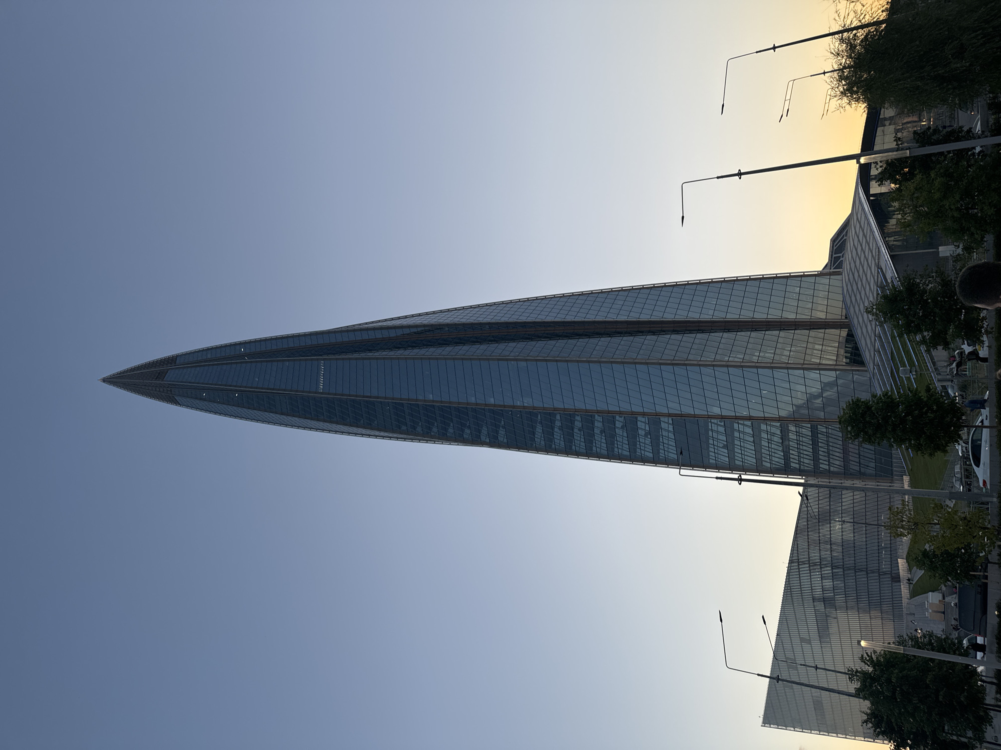 An ultra-wide shot captures the Lakhta Center skyscraper in Sinyavino, Russia, during the golden hour. The towering glass structure dominates the frame, its pointed apex reaching towards a clear, pale blue sky that transitions to a soft yellow-orange hue near the horizon. The building's modern design features a sleek, curved facade composed of countless glass panels, reflecting the ambient light. To the left of the skyscraper, a lower, angular building with a similarly glass-covered exterior is visible, along with a sloping green area and a series of tiered walkways. The foreground reveals streetlights with modern, minimalist designs, and a scattering of trees with lush green foliage. Several cars are parked or driving in the middle ground, and the silhouettes of a few individuals can be seen walking or standing near the buildings. The overall scene conveys a sense of contemporary urban architecture set against a serene twilight sky.