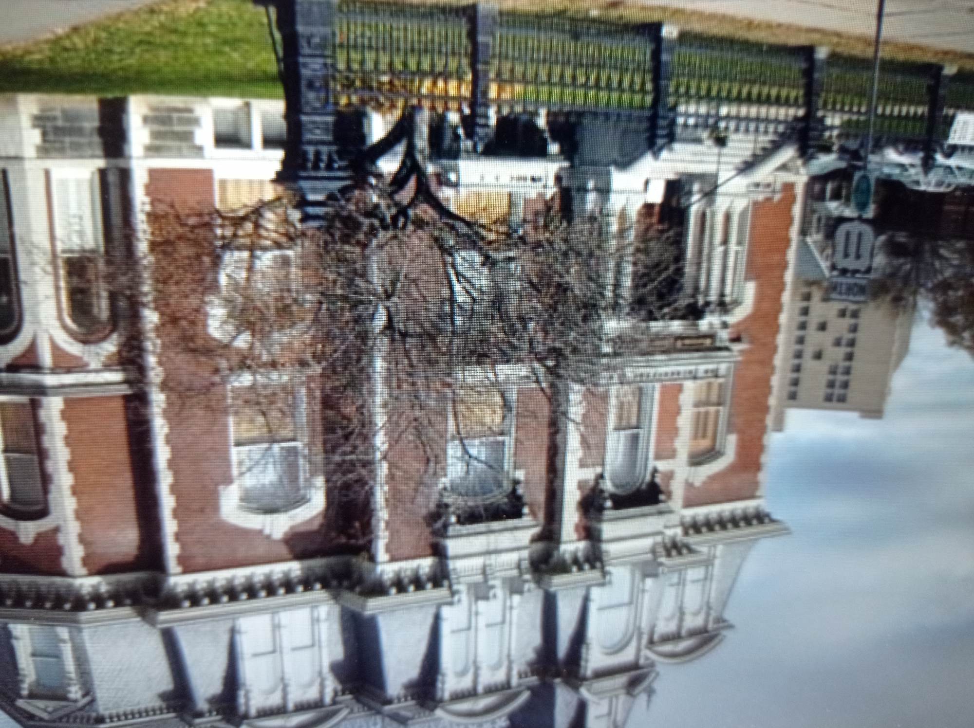 A large, multi-story brick building with ornate architectural details stands prominently in the foreground. The building features a mansard roof with dormer windows, decorative cornices, and arched windows. A tree with bare branches is situated in front of the building, partially obscuring the view. A black metal fence runs across the front of the property. To the left of the brick building, a taller, modern building with many windows is visible. A street sign indicating "NORTH 11" is also present on the left side of the image. The ground in front of the fence is covered with green grass. Cars are parked on a street in the middle ground. The sky is overcast. The image is a photograph taken from a slightly low angle.
