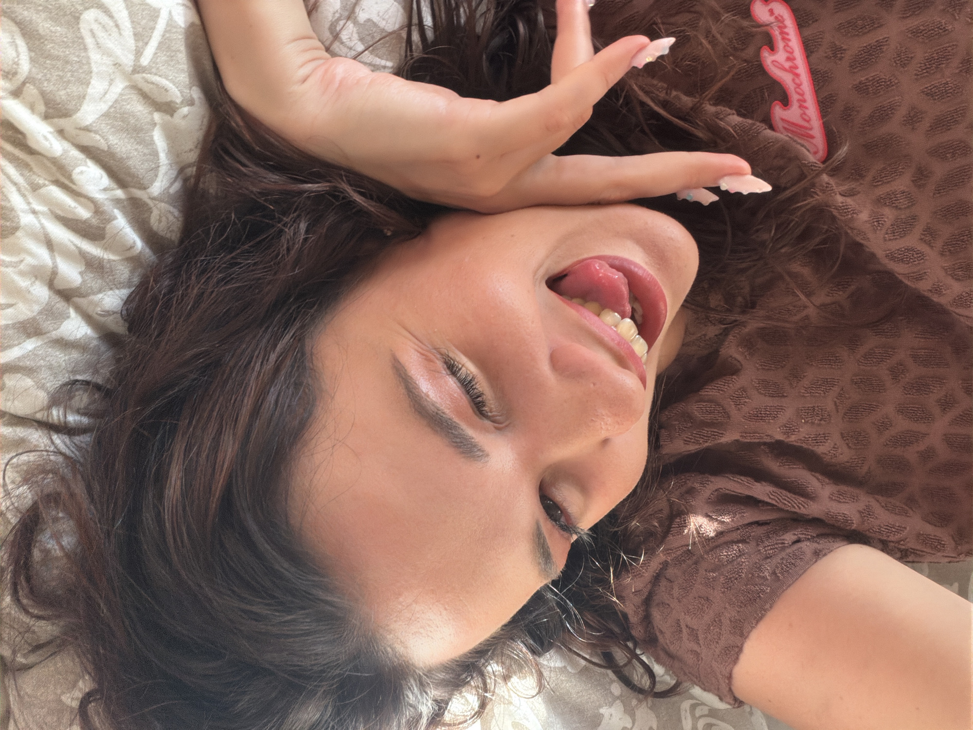 A young woman is lying on her back on a bed with patterned sheets, her head tilted to the side. Her eyes are closed, and her mouth is open, with her tongue sticking out. Her right hand is raised to her face, with her fingers spread. She is wearing a brown, textured t-shirt with a pink "Monochrome" logo on the left side. The lighting suggests it is daytime, with sunlight casting shadows on her face and hair. The location is not specified in the image.