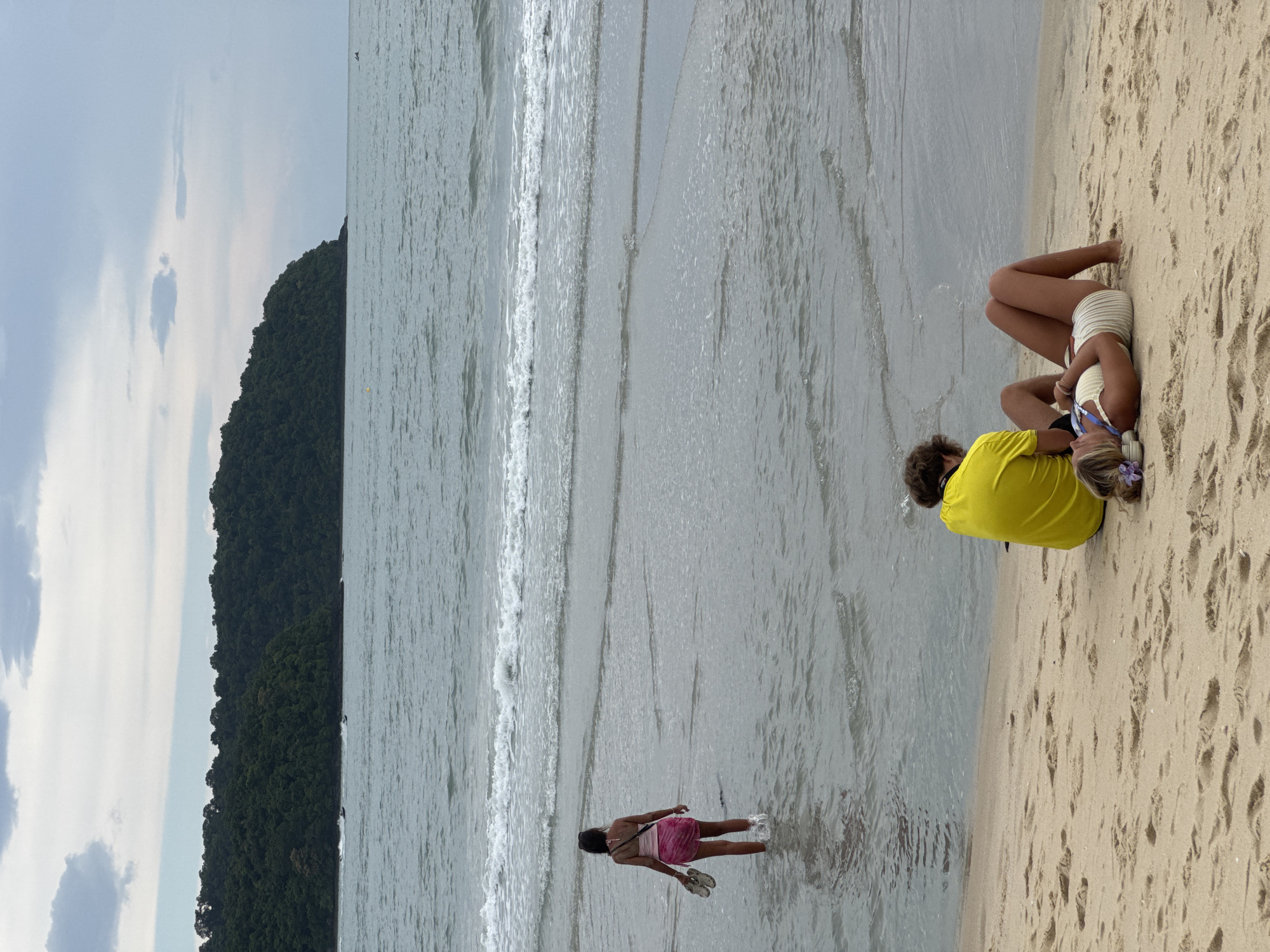 The media depicts a beach scene on Tengah Island, Malaysia, with two people on the sand and one wading in the shallow water. The sky is overcast with light clouds, suggesting it's daytime, possibly late afternoon. The sea is calm with gentle waves lapping the shore.
On the sand, closer to the camera, a person wearing a bright yellow shirt is seated, facing away from the viewer. Next to them, another person lies on their side, wearing a white textured outfit. They appear relaxed.
Further into the water, a person in a pink dress is standing, with their feet submerged in the sea. They are holding a pair of sandals. The overall mood seems peaceful and relaxed, indicative of a casual day at the beach.
A lush, tree-covered hillside forms the backdrop, suggesting the island's natural beauty. The sand is marked with footprints, indicating recent activity on the beach. No visible text is present in the image.