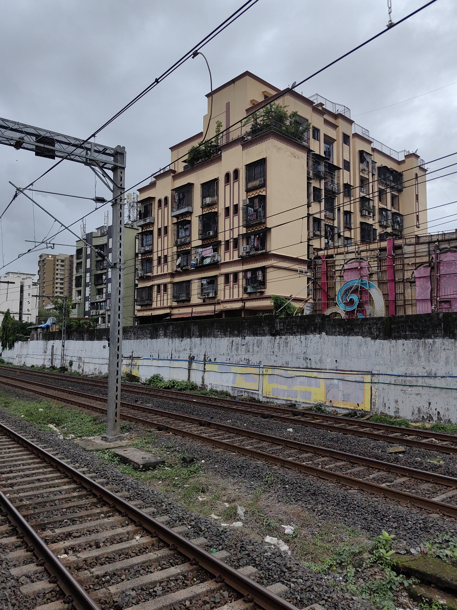 This is a daytime, eye-level shot of a railway track with an urban background in Mumbai, India. The scene is dominated by the overcast sky and the stark lines of the railway infrastructure juxtaposed with residential buildings.
In the foreground, multiple sets of railway tracks recede into the distance, their gravel ballast interspersed with patches of green grass and some scattered debris. A tall, grey metal pole, part of the overhead electrical system for the trains, stands prominently between the tracks.
Behind the tracks, a weathered concrete wall runs horizontally across the frame. It appears to have some faded painted murals or graffiti on it, with yellow and blue sections visible. Above the wall, a cream-colored, multi-story apartment building is the most significant structure. It features numerous balconies with ornate metal grills, many with clothes hanging out to dry. The building has a flat roof with some greenery and railings. To the left of this building, other apartment buildings of varying heights and architectural styles are visible, including a taller, lighter-colored building with the word "CARROT" visible in its upper floors. Further left, a cluster of smaller, darker buildings is present.
To the right of the cream-colored building, behind the wall, there is a vibrant, large-scale, abstract art installation or advertisement. It consists of bright pink panels and a large, swirling blue and white spiral design. The installation appears to be supported by a metal framework.
The overall atmosphere is somewhat muted due to the cloudy weather. No people are clearly visible in the frame, and no specific actions are being depicted. The image captures a typical view of urban infrastructure and residential living along a railway line in Mumbai.