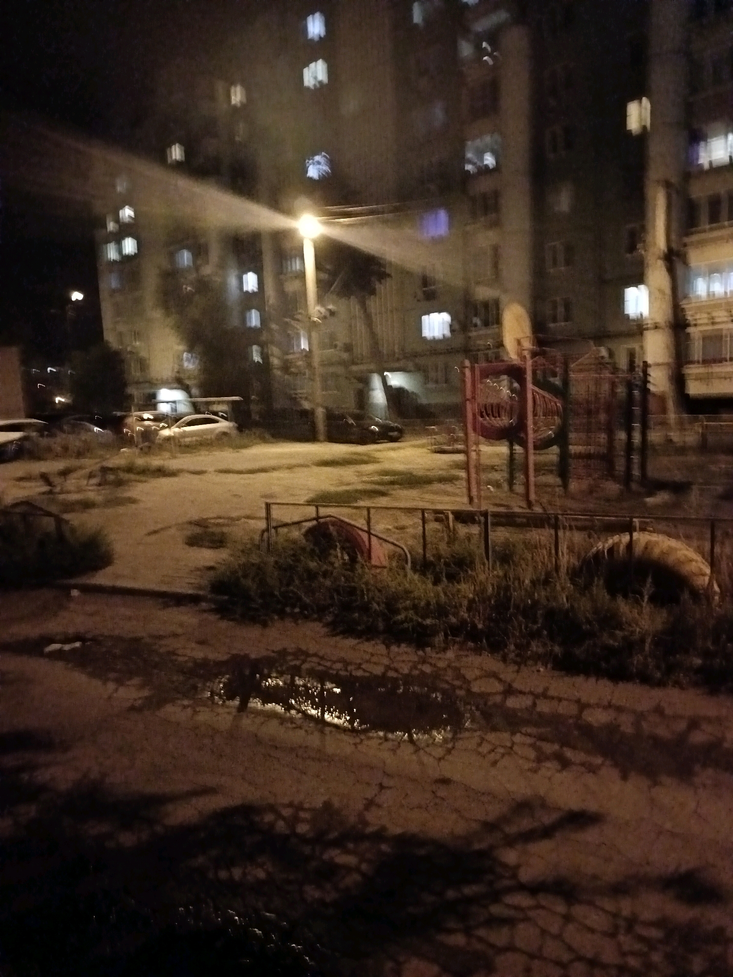 The image depicts an outdoor scene at night, likely in a residential area of Astrakhan, Russia. A multi-story apartment building is visible in the background with numerous windows illuminated, indicating occupancy. In the foreground, there is a playground with red and blue structures, including slides and climbing frames. Adjacent to the playground is a patch of overgrown grass and a fence. The ground surface appears to be a mixture of dirt and asphalt, with several puddles reflecting the ambient light. Several cars are parked in the vicinity, with at least one white sedan clearly visible. Shadows from nearby trees and possibly streetlights are cast across the ground, creating a somewhat eerie atmosphere. There are no people clearly visible in the image. The overall impression is of a typical urban setting after dark, with residential buildings and recreational facilities.