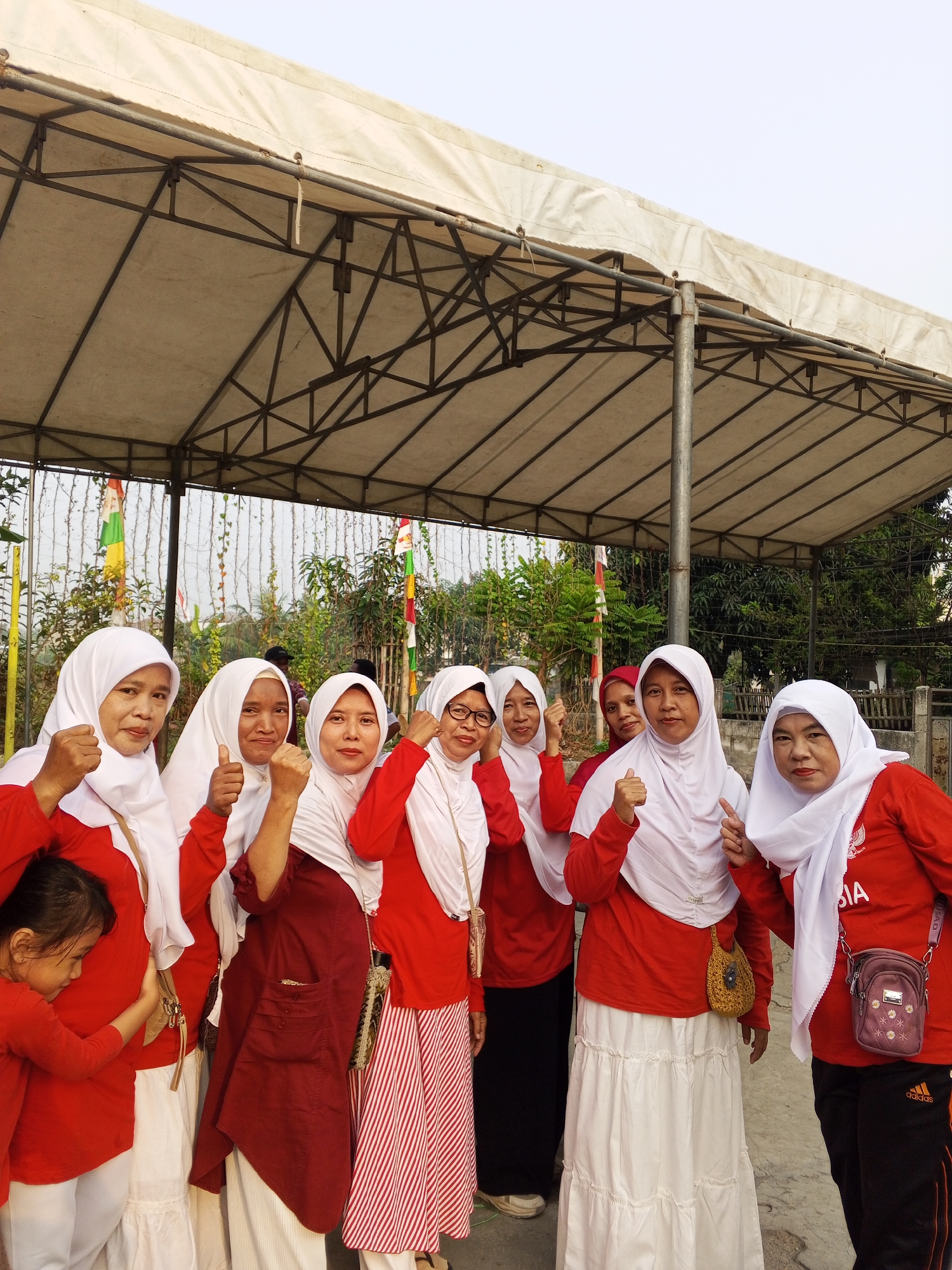 Eight individuals, seven women and one young girl, are posing for a photo under a large, white, canvas-covered canopy with a metal frame. The canopy is partially supported by a thick vertical metal pole. The women are wearing red tops and white headscarves, with some also wearing white or striped skirts, and one woman in black pants with an Adidas logo. The young girl is wearing a red top and white pants. They are all facing forward, with some of the women making fist gestures, and one giving a thumbs-up. In the background, there are trees and foliage, and several Indonesian flags are visible, suggesting a celebratory or national event. The ground appears to be paved. The location is identified as Ciruas, Indonesia.