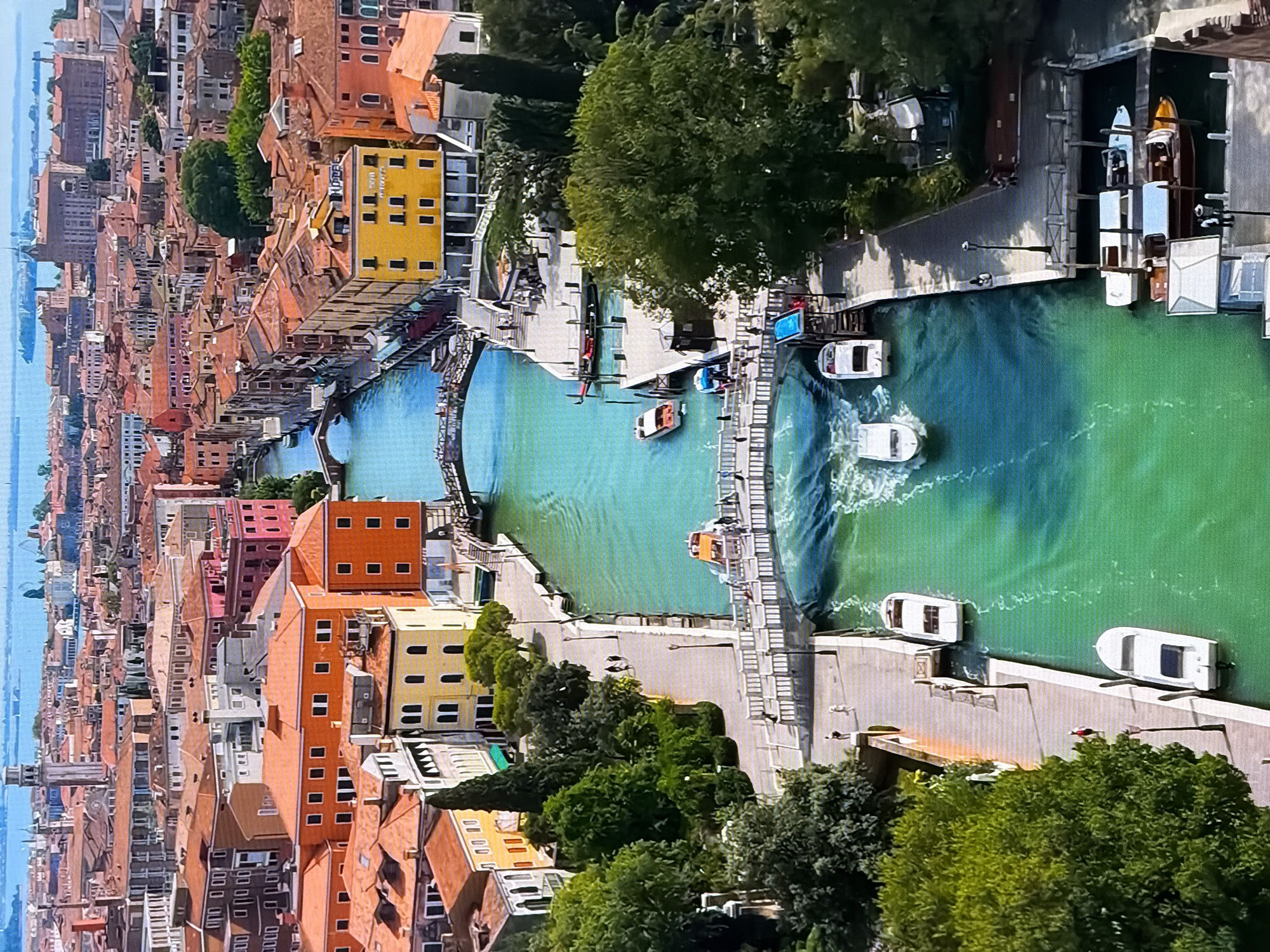 This is an aerial, high-angle, wide shot of a canal in Venice, Italy. The canal is a vibrant turquoise color and is filled with several boats, including speedboats, a water taxi, and what appears to be a gondola. The canal is lined with tall, narrow buildings with red-tiled roofs, characteristic of Venetian architecture. There are several bridges crossing the canal, with people walking across them. The foreground of the image shows a lush green tree and a paved walkway. The background reveals a cityscape with more buildings and a distant body of water. The scene is bustling with activity, with boats navigating the canal and people moving along the walkways.
The prompt requests information about Chirchik, Uzbekistan, but the image clearly depicts Venice, Italy. Therefore, I cannot fulfill the request to describe the location in Chirchik, Uzbekistan.