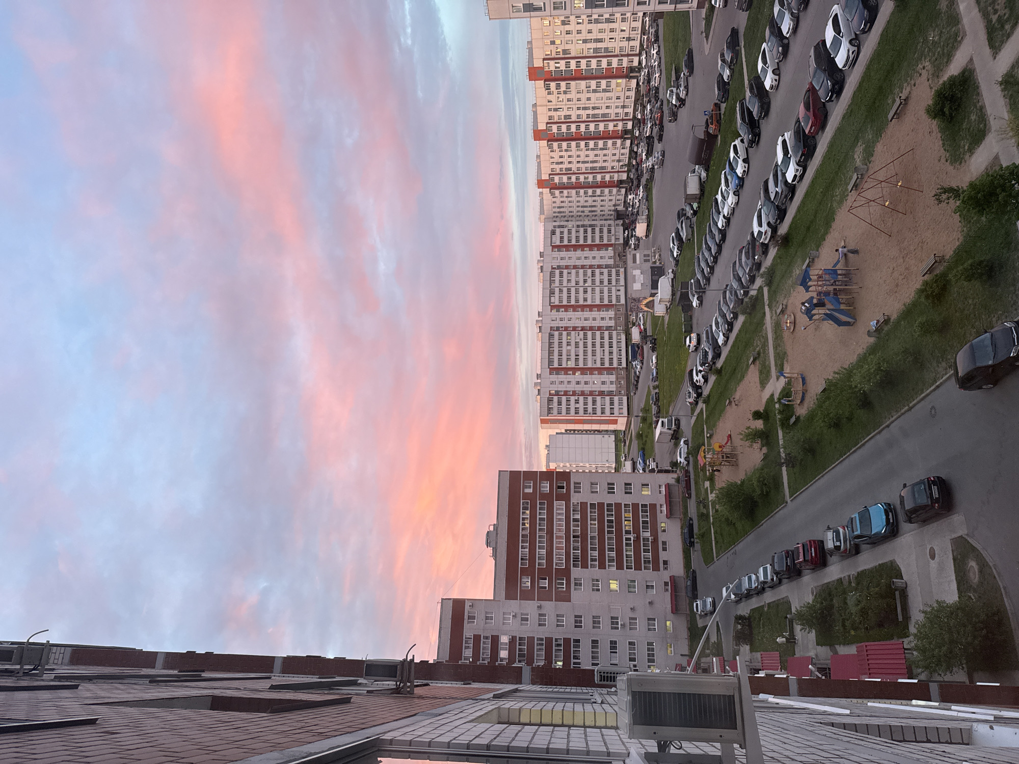 An elevated, oblique view shows a residential neighborhood during twilight, likely in Veliky Novgorod, Russia, though there is no direct evidence to prove it. The sky is striated with pink and blue hues. Several high-rise apartment buildings of similar design with white and red facades are visible. Long rows of parked cars line the streets and sidewalks next to the buildings. A playground with swings and slides is situated in a sandy area adjacent to the parking. A building rooftop with a reddish-brown tiled surface is visible at the very bottom of the image, suggesting the photograph was taken from a high vantage point. There is no indication of people in the image.