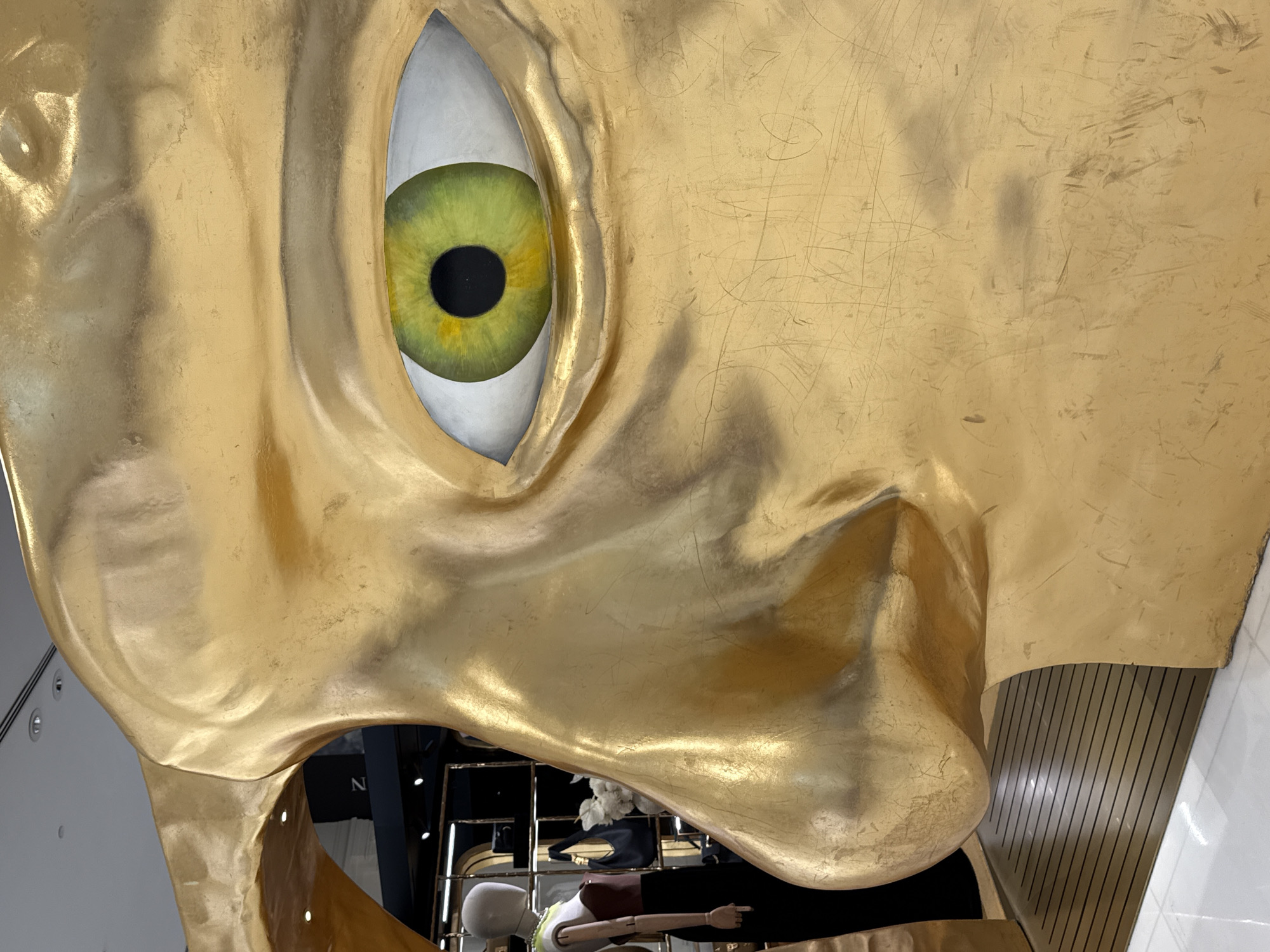 The image shows a close-up of a large, golden, abstract sculpture of a face, possibly a stylized representation of a human nose and eye. The sculpture is located in a commercial setting, likely a retail store or mall, in Dubai, United Arab Emirates. The background is blurred, but hints of mannequins dressed in clothing, display shelves, and store lighting are visible. The sculpture has a metallic, golden finish and shows signs of wear, such as scratches and smudges. The eye of the sculpture is painted with a vibrant green iris and a black pupil. The lighting suggests an indoor environment with artificial illumination.