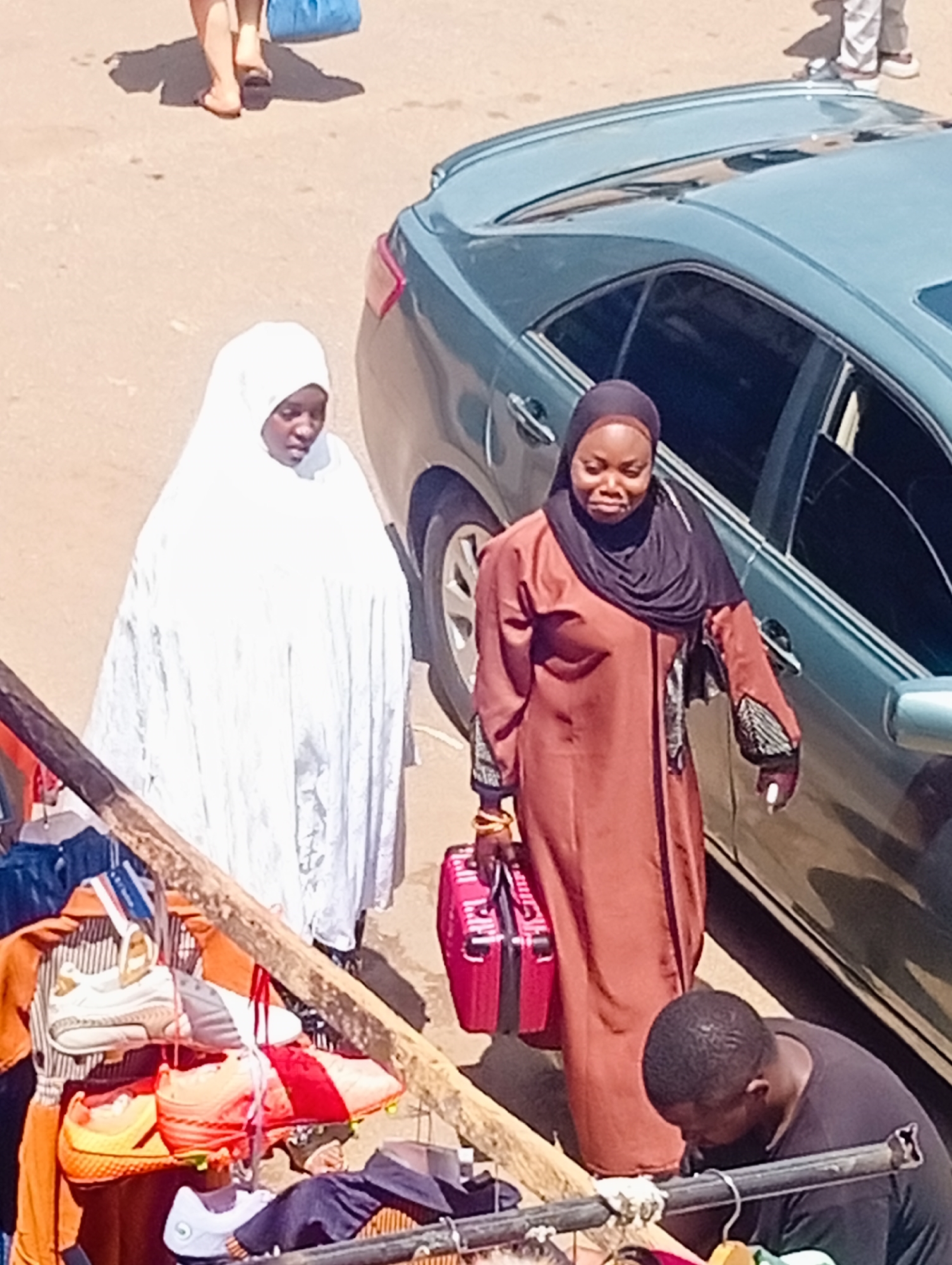 Two women and a man are in a market scene in Bukuru, Nigeria. The woman on the left is wearing a white hijab and a long white garment. The woman on the right is wearing a black hijab and a rust-colored long dress. She is holding a red suitcase. The man in the foreground is wearing a black t-shirt and is bent over looking at items on a display rack. In front of the man is a rack with several pairs of shoes and other clothing items hanging. A light blue car is parked to the right of the women. The lighting suggests it is daytime, likely with bright sunlight creating shadows. The overall scene depicts a busy marketplace with vendors and shoppers. No visible text is present in the image.