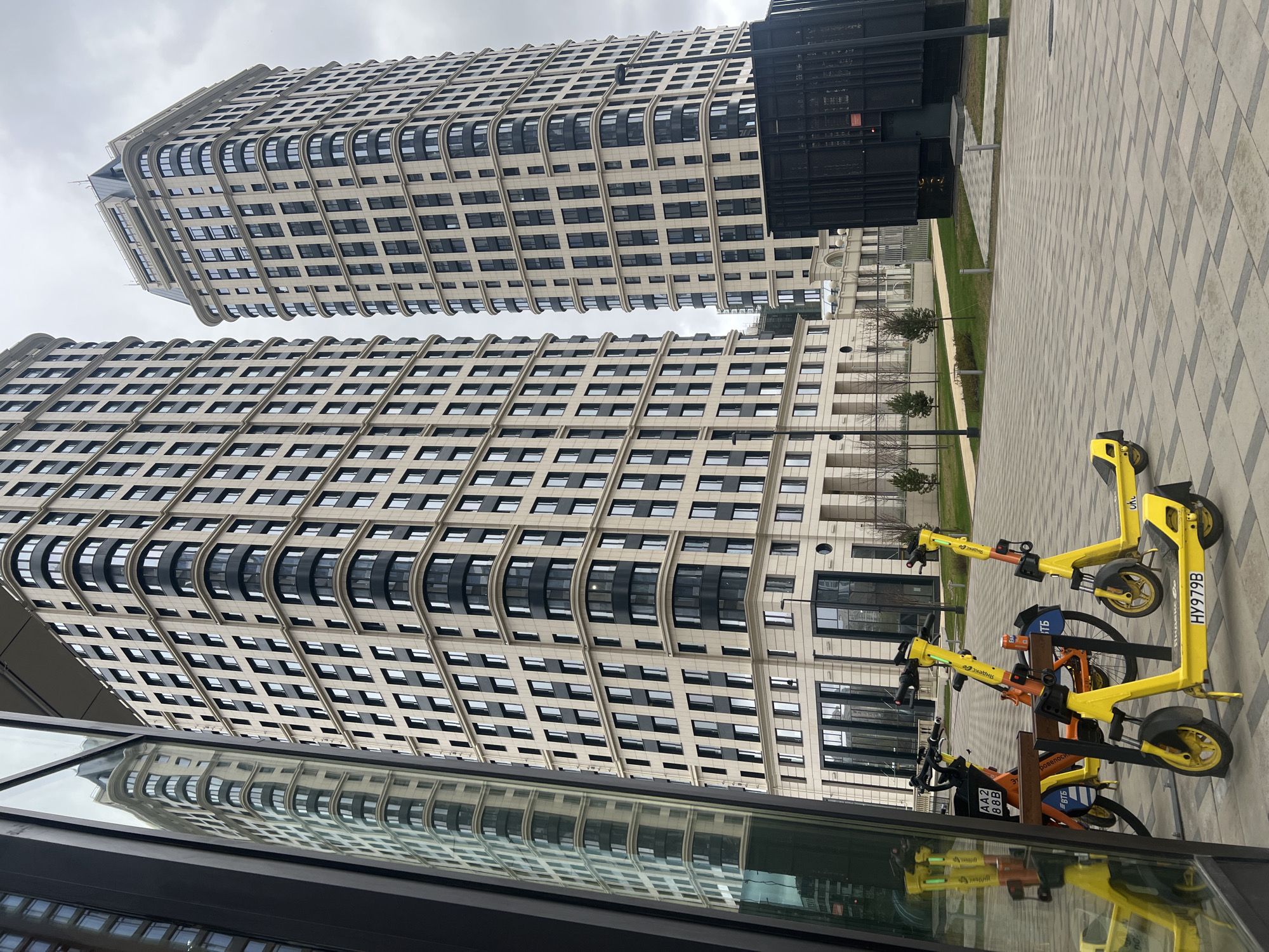 Two tall, modern buildings dominate the view. Their architecture features a repeating grid pattern of windows, accentuated by curved sections. A black rectangular structure sits adjacent to one building. In the foreground, several brightly colored yellow scooters stand parked on a paved area. The buildings' reflections are visible in a glassy surface at the bottom left corner. The sky is overcast. The location is in the city of Khimki, Russia.