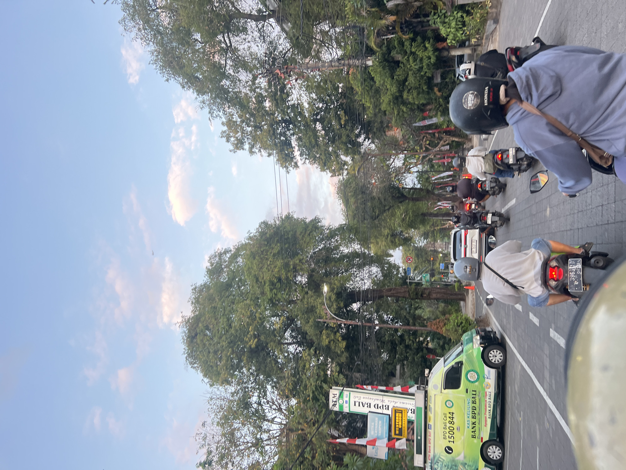 In Denpasar, Indonesia, a street scene unfolds with multiple people riding motorcycles. The perspective is from behind one of the riders, looking forward down a road lined with trees and various signs. On the left side of the road, a green and white van with "BANK BPD BALI" branding is visible, along with a sign for "ATM BPD BALI" and "WESTERN UNION". Several motorcycles are proceeding in the same direction as the viewer, some with passengers. A white car is also in traffic ahead. The sky above is a light blue with scattered clouds, suggesting it is late afternoon or early evening. The overall impression is of a busy urban street with typical traffic and commercial signage.