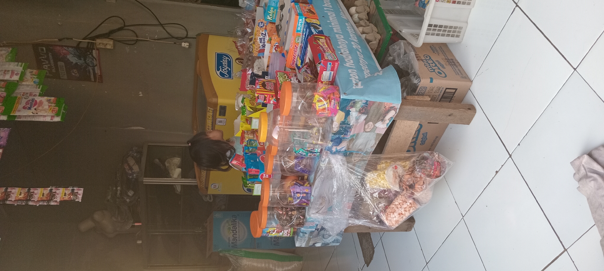 In Mataram, Indonesia, a young girl with dark hair pulled into a ponytail stands behind a table laden with an assortment of snacks and drinks. She is partially obscured by the items on display. The table is covered with a light blue cloth adorned with colorful illustrations. Arranged on the table are various plastic containers filled with individually wrapped candies and snacks, as well as some boxed items. Behind the table, a yellow refrigerator with the "Joyday" logo is visible, likely used for chilling beverages. To the left, a large blue box labeled "Mandalika" and a white sack are present. The overall environment appears to be a small shop or kiosk, with shelves and merchandise visible in the background, suggesting a commercial setting. The floor is tiled in a grid pattern.