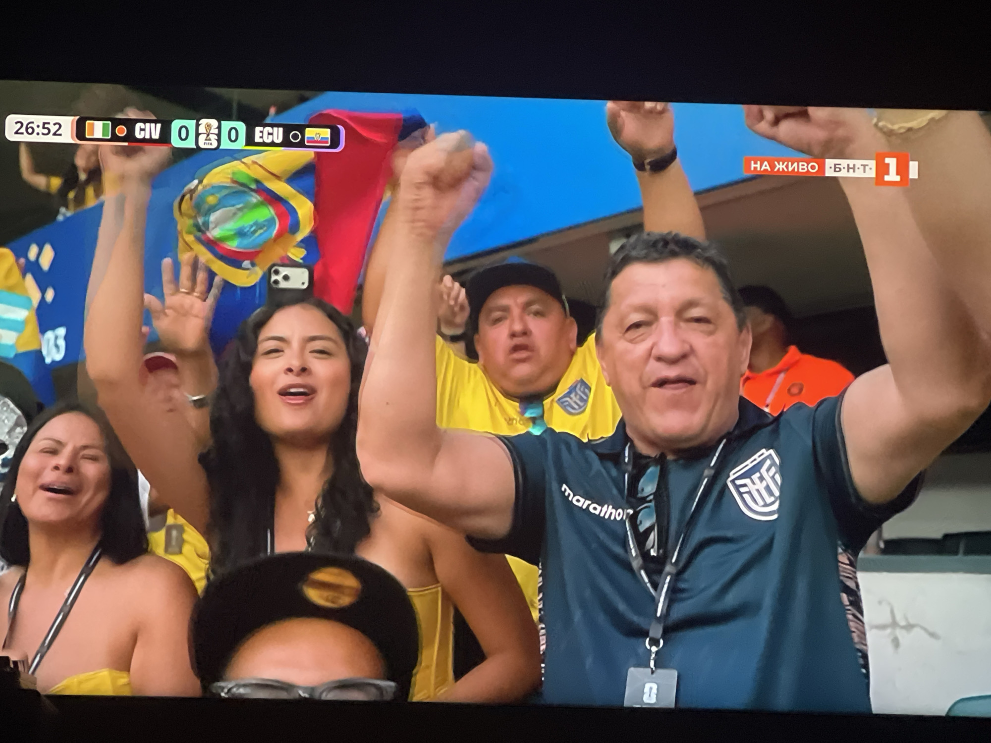 The image depicts a screen displaying a soccer match score with "CIV 0" and "ECU 0" at 26:52. The background shows a crowd of enthusiastic soccer fans, predominantly wearing yellow jerseys, waving flags and cheering. A woman in the foreground on the left is smiling widely, her mouth open in a cheer. Beside her, a man in a dark blue shirt with a "marathon" logo and an Ecuadorian flag on his sleeve is raising his fist in celebration. To the right, a man in a bright yellow shirt is also cheering. The screen also shows "НА ЖИВО Б·Н·Т 1" indicating a live broadcast on a Bulgarian television channel. The overall scene captures the excitement and energy of a live sporting event.