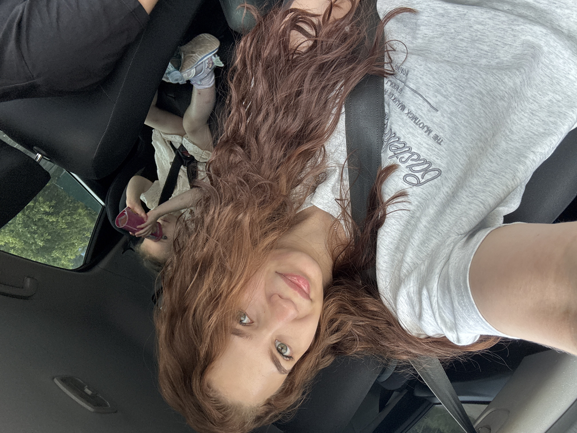 A selfie-style photo taken inside a car. A young woman with reddish-brown wavy hair and fair skin is positioned in the lower center of the frame, facing the camera. She has green eyes. She is wearing a gray t-shirt and is secured with a seatbelt. Behind her, in the upper left corner of the frame, is a child wearing a white top with lace details, black pants, and white and blue shoes. The child is holding a pink cup and appears to be drinking from it. The car's interior is predominantly black. The car's window is visible in the upper left, showing greenery outside, potentially indicating a park or treed area. Based on the prompt, the location is Chaikovsky, Russia.