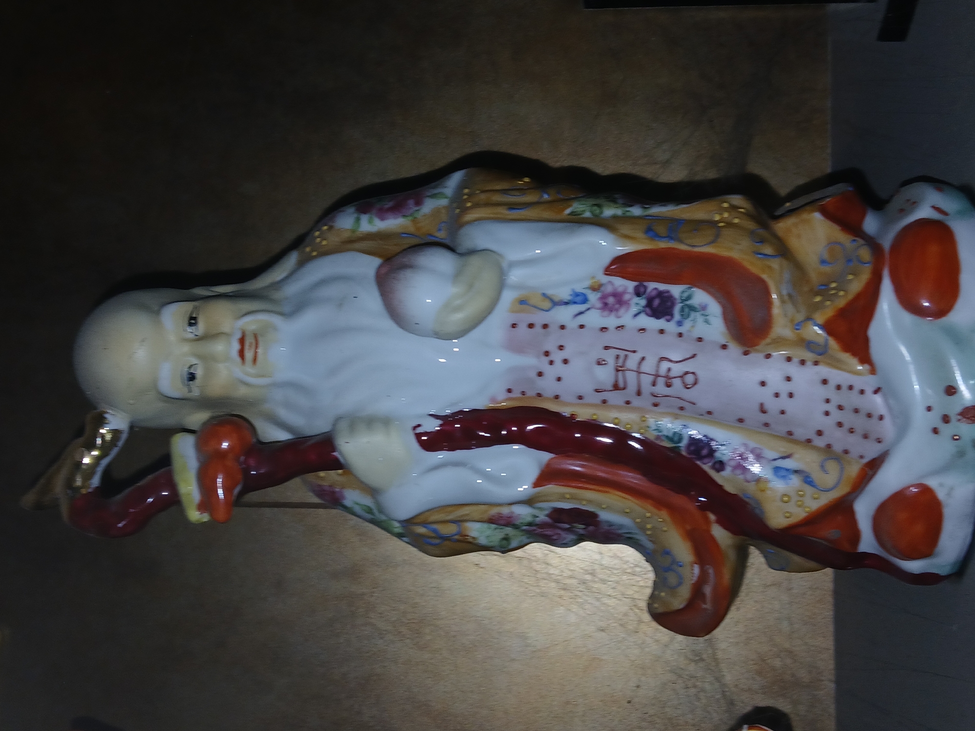 The media file displays a ceramic or porcelain figurine of an elderly man, likely a Chinese deity such as Shou Xing (God of Longevity), lying on his back. The figure has a large bald head, a long white beard, and a serene, benevolent expression with dark eyes and red lips. He wears an ornate, richly colored robe primarily in white, gold, and red/orange. The robe is decorated with intricate floral patterns in shades of pink, purple, blue, and green, along with small yellow and blue swirl motifs, and numerous small red dots.
The figurine holds a red staff with yellow accents in his right hand. A prominent, peach-shaped fruit, possibly a peach of immortality, rests on his chest/belly. A golden earring or ring is visible on his left ear. On the white section of his robe, above the peach, there are reddish-brown stylized Chinese characters, one of which appears to be "壽" (shòu), meaning longevity.
The figurine is positioned on a flat, light brown or tan surface, possibly a wooden table. The lighting is somewhat dim, creating soft shadows around the object. No specific activity is depicted beyond the static pose of the figurine. The overall aesthetic is traditional East Asian, showcasing detailed craftsmanship and vibrant colors.