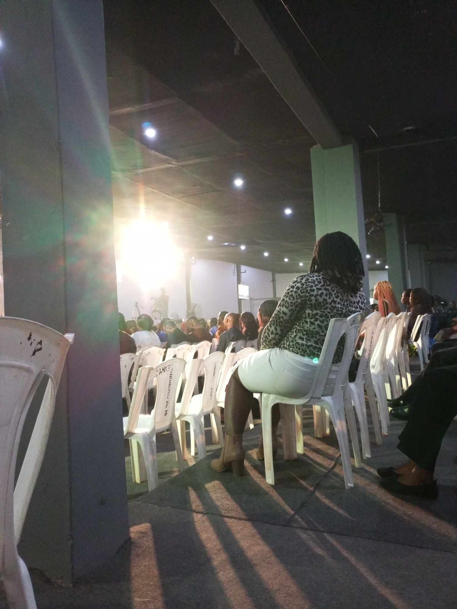 The image depicts an indoor event space, likely a hall or auditorium, in Jos, Nigeria. The scene is illuminated by bright, overhead lights, one of which creates a strong lens flare in the upper left quadrant. The ceiling is dark, with visible beams and lighting fixtures. In the foreground, a woman with braided hair and a leopard-print top is seated in a white plastic chair, facing away from the camera. She is wearing a white skirt and heeled boots. Rows of similar white plastic chairs are arranged throughout the space, some occupied by other individuals whose faces are not clearly visible due to distance or orientation. The floor appears to be paved, with distinct shadows cast by the chairs and the lighting. A large, dark pillar is prominent on the left side of the image, partially obscuring the view of the seated woman. The overall atmosphere suggests a gathering or a performance.