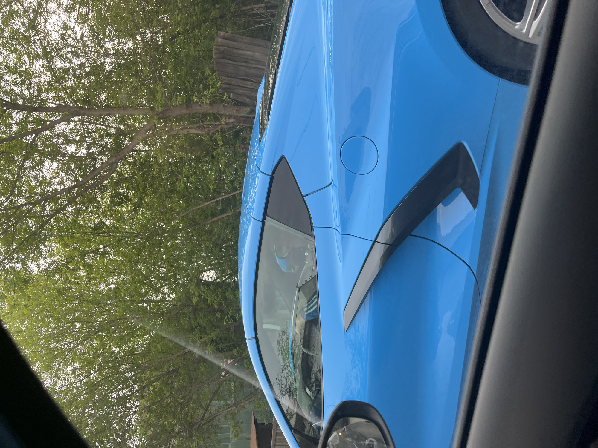 A side view of a light blue car is partially visible in a photograph taken from inside another vehicle. The car has black side vents and a black trim around the windshield. Trees are visible in the background. The location is not discernible from the image.