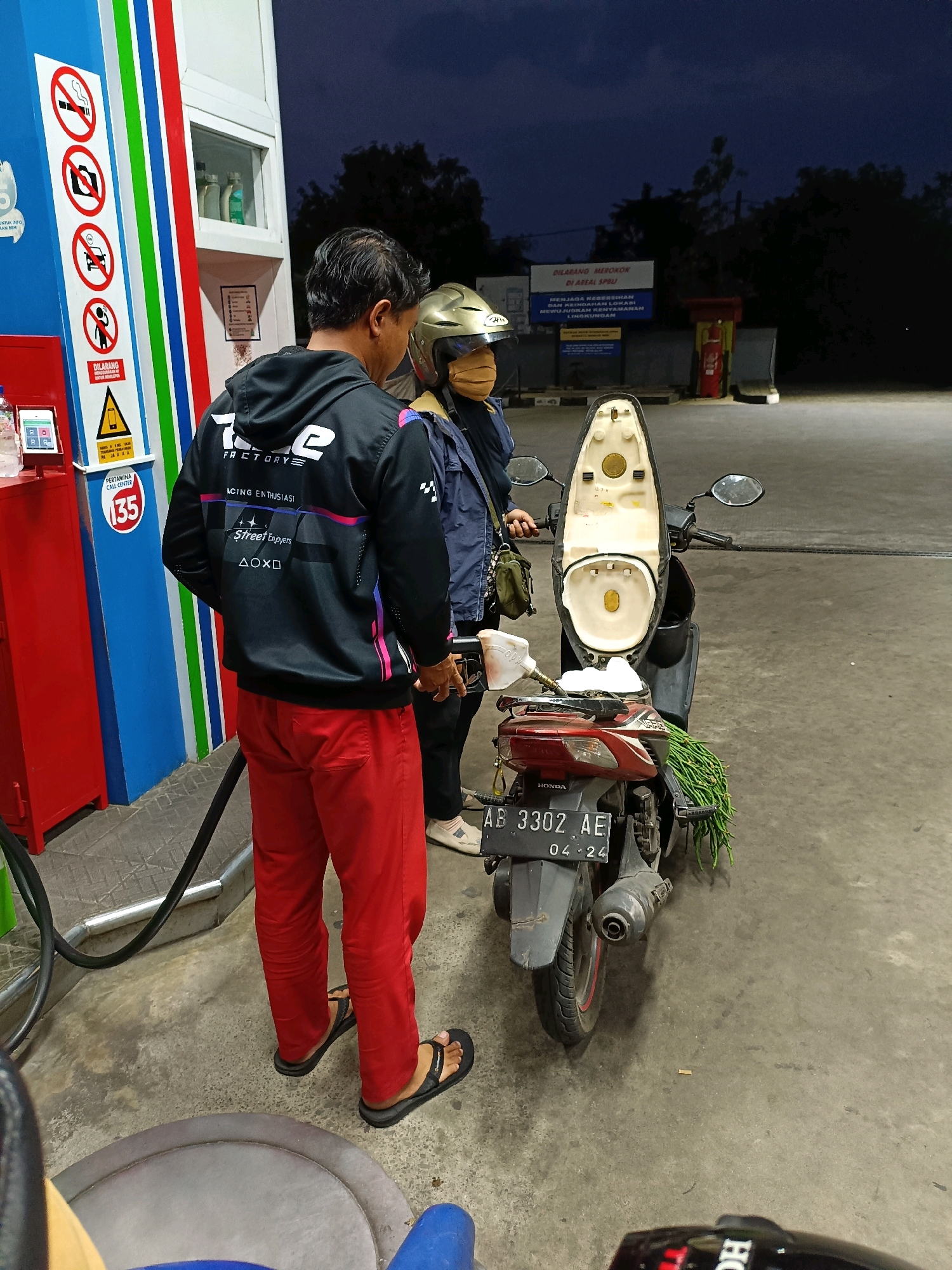 At a gas station in Maguwoharjo, Indonesia, at night, a man wearing red pants and a black hooded jacket with white and pink accents is standing next to a scooter. He appears to be fueling the scooter, as he is holding a gas nozzle. A woman, wearing a blue jacket and a gold helmet with a light brown face mask, stands behind him, holding a green bunch of produce. The scooter has a license plate that reads "AB 3302 AE 04-24". In the background, there are gas pumps and signage. The ground is paved, and there is vegetation visible in the distance.