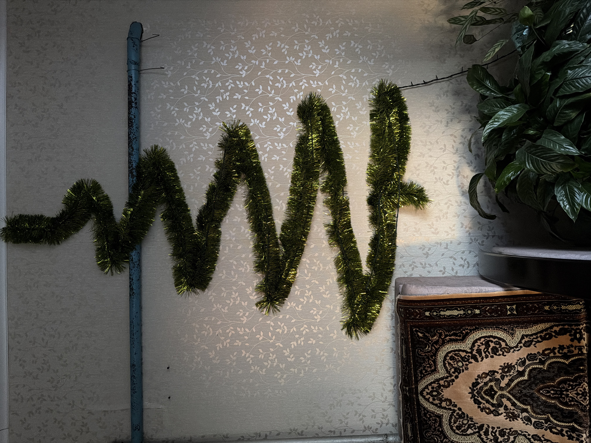 The image shows an interior scene in what appears to be a residential setting in Кадряково, Russia. The wall is decorated with a repeating leaf pattern wallpaper. A length of green tinsel is attached to the wall, forming a wavy, somewhat erratic line. To the left of the tinsel, a light blue pipe runs vertically. To the right, a dark green leafy plant is visible in a black pot sitting on a light colored stool or cushion. A patterned rug or fabric is draped over the stool or cushion. The lighting is somewhat uneven, with shadows present on the left side of the image. The scene suggests a space possibly decorated for a holiday or other festive occasion.