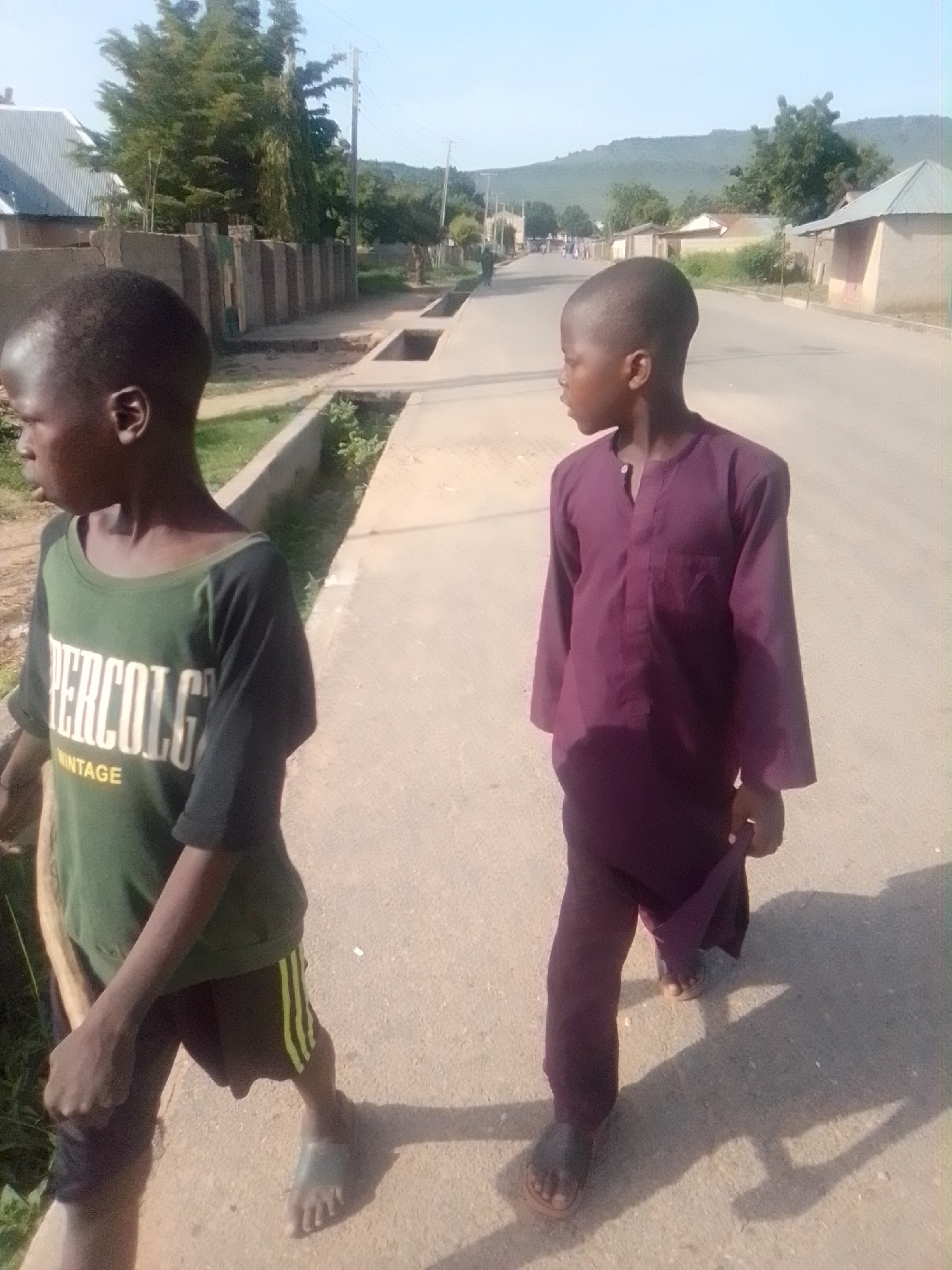 The image depicts two young boys walking on a paved road in Talasse, Nigeria. The boy on the left wears a green t-shirt with white lettering that reads "PERCOLG VINTAGE" and black shorts with yellow stripes. The boy on the right is wearing a maroon, long-sleeved tunic and dark pants. Both boys are wearing sandals. The road is lined with a concrete drainage ditch on the left and a low concrete wall with vertical posts. In the background, there are buildings with corrugated metal roofs, trees, and a gently sloping hill under a clear blue sky. The lighting suggests it is daytime with sunlight casting shadows.