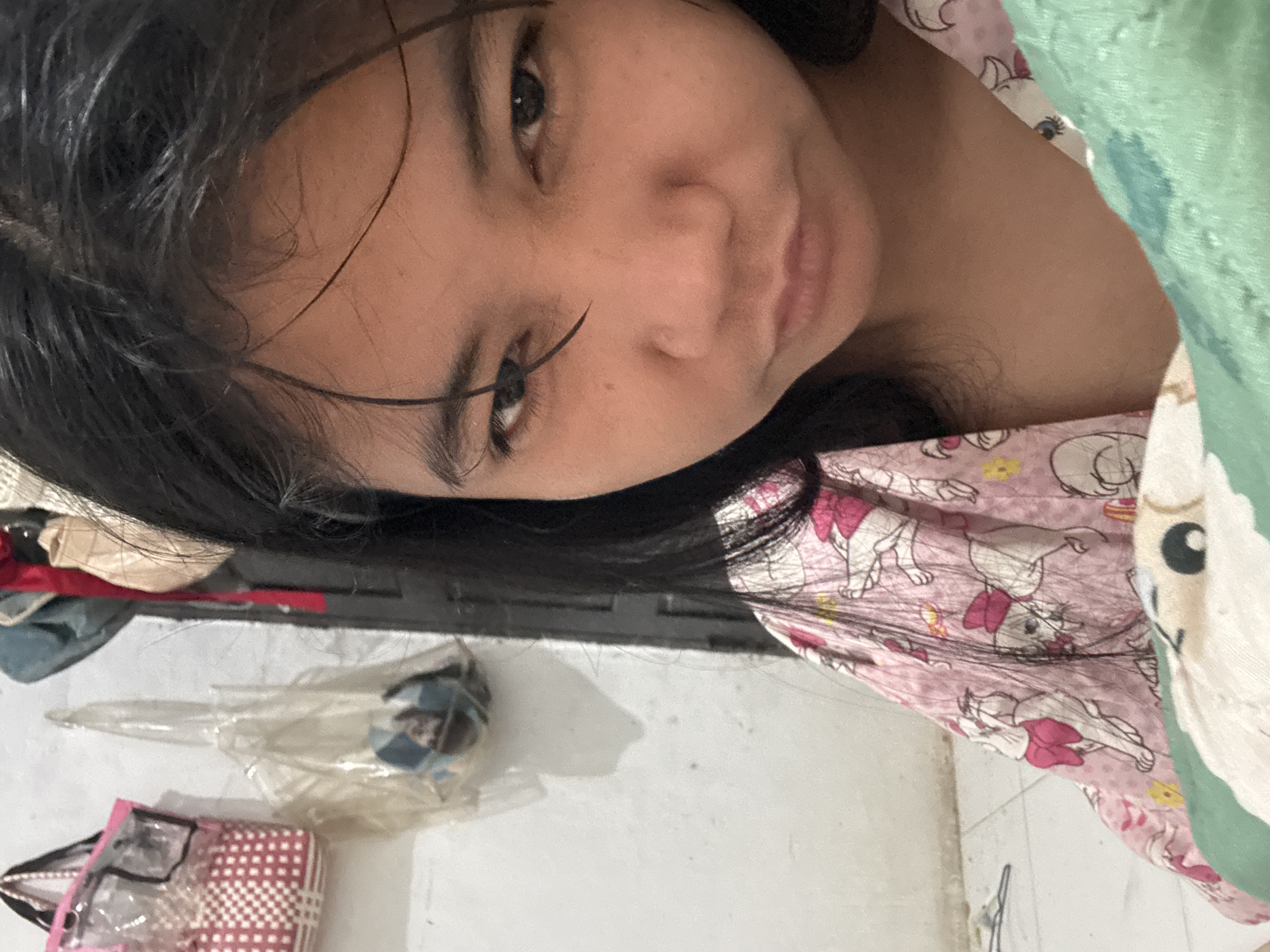 The image is a close-up selfie of a young woman with long, dark, slightly messy hair and dark eyes. She is wearing a pink pajama top with a cartoon character print. Her expression is somewhat pouty. The background shows a white wall with some items hanging on it, including a pink and white checkered bag and some clear plastic bags containing what appear to be other objects. To the right, a dark vertical surface, possibly a door or cabinet, is visible. A portion of a green and white patterned blanket or comforter is in the foreground. The scene appears to be indoors, likely a bedroom. There is no information in the image to suggest it is specifically located in North Jakarta, Indonesia.