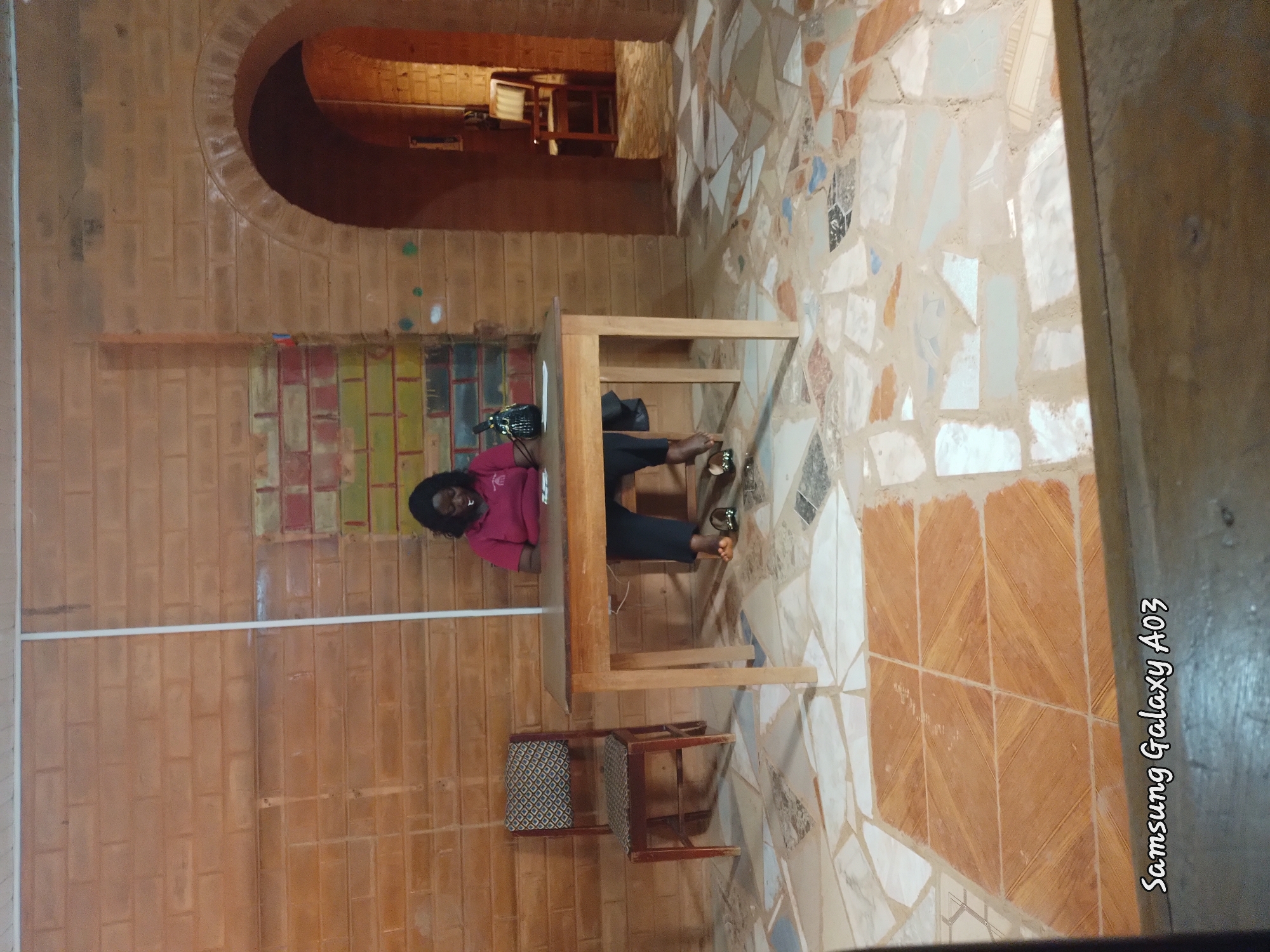 The image shows a woman sitting at a wooden table in a room with brick walls and a tiled floor. She is wearing a pink shirt and black pants. The floor is decorated with an abstract mosaic pattern of various colored tiles. A wooden chair with a patterned seat is placed in front of the table. The room appears to be indoors, with a large arched opening in the brick wall revealing more of the interior. The lighting suggests it might be daytime, or the room is well-lit. The woman is looking towards the camera and smiling, appearing relaxed. The overall atmosphere seems casual and inviting. The text "Samsung Galaxy A03" is visible along the right edge of the image, indicating the device used to capture the photo.