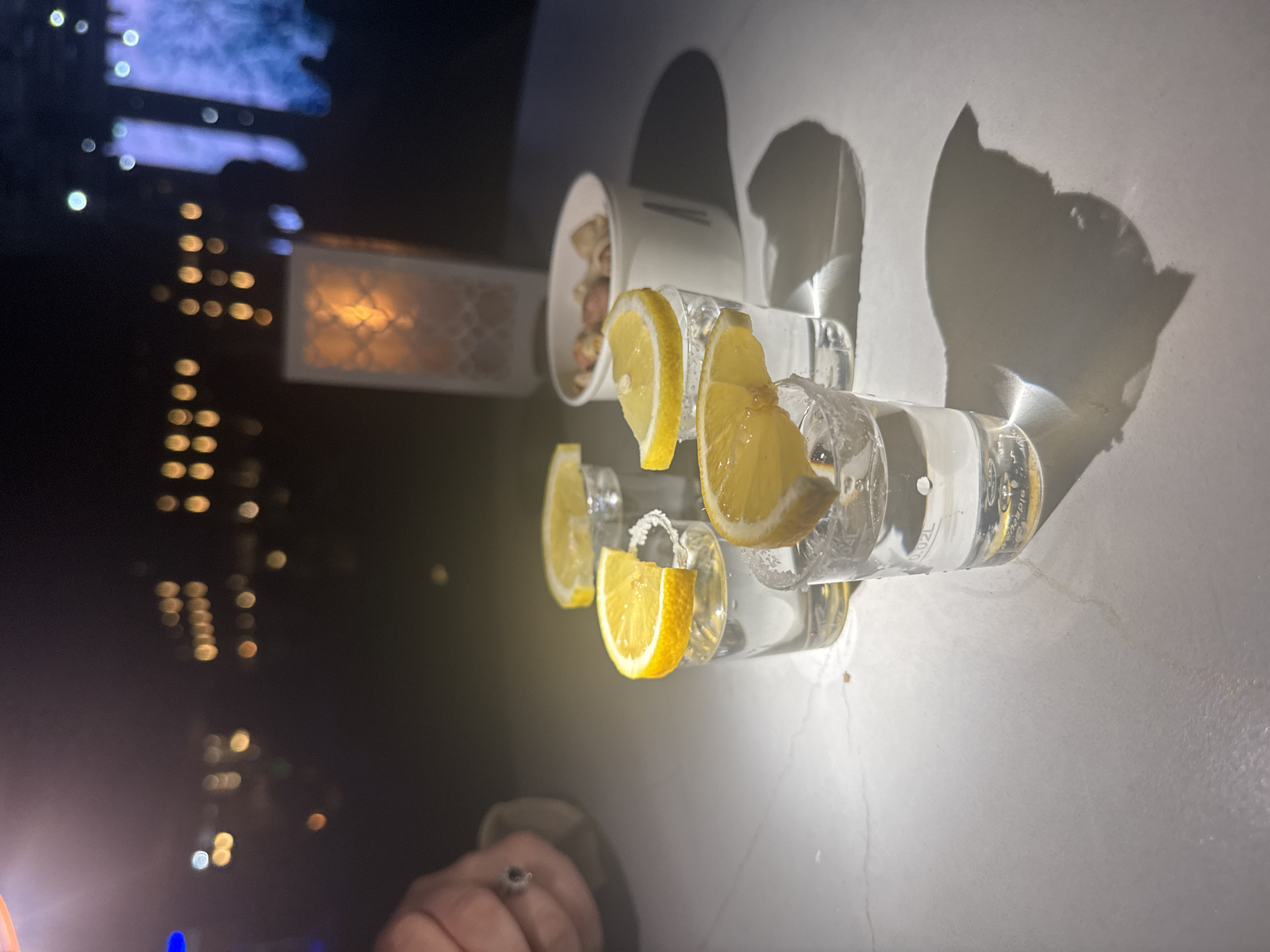 The image depicts a close-up, eye-level view of four shot glasses filled with a clear liquid, likely a spirit. Each glass is garnished with a slice of lemon, some of which are perched on the rim, coated with salt. The shot glasses are arranged on a white, textured surface, possibly a table, with shadows cast by the glasses and their contents. In the background, out of focus, are the lights of buildings and possibly a body of water, suggesting an outdoor setting at night. A white paper container filled with nuts is also visible in the mid-ground, as well as a decorative lantern. A hand, adorned with a ring, is partially visible on the left side of the frame. The overall scene suggests a bar or restaurant setting, potentially in a resort area. The environment appears to be Manavgat, Turkey, as inferred from the provided context.