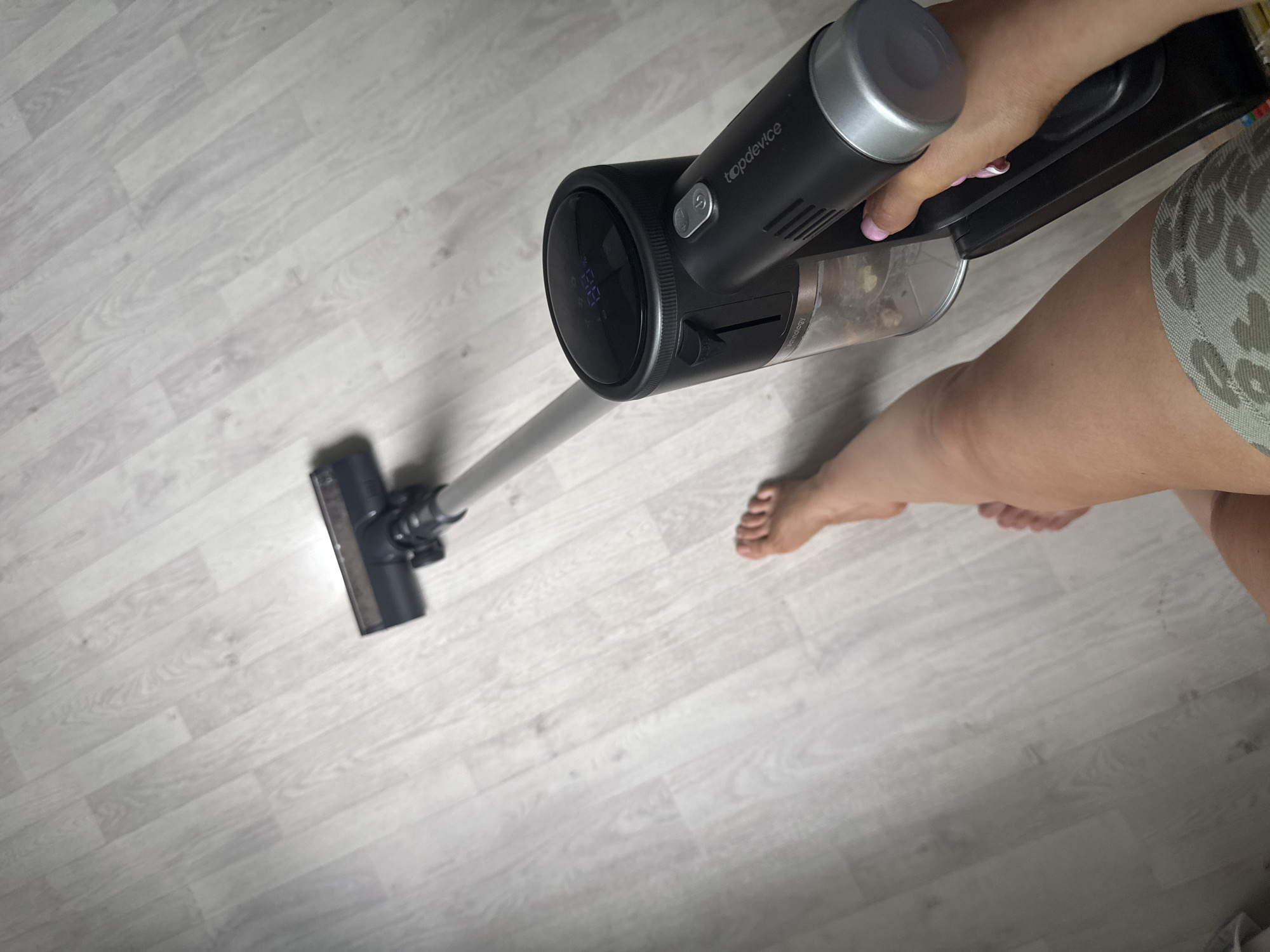 A first-person perspective shot shows a person vacuuming a light-colored laminate floor. The vacuum cleaner, a black stick vacuum with a transparent dustbin showing debris, is held in the person's right hand. The vacuum's digital display shows "88". The person's bare feet are visible on the floor to the left of the vacuum, with their legs and patterned shorts visible in the lower right corner. The scene appears to be indoors, likely in a residential setting. There is no discernible indication of the specific city or country.
