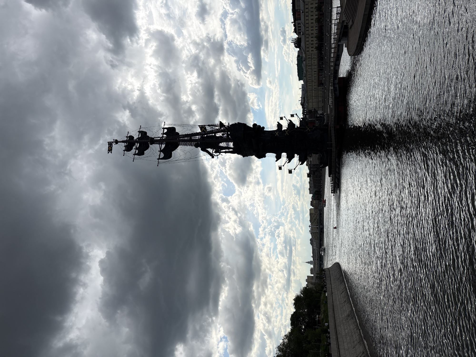 The image shows a silhouette of a tall ship statue against a dramatic cloudy sky, reflected in the dark water. In the background, the architecture of Moscow is visible along the riverbank. The statue's dark silhouette stands out against the bright sky, creating a contrast. A retaining wall is visible in the foreground on the right side of the photo.