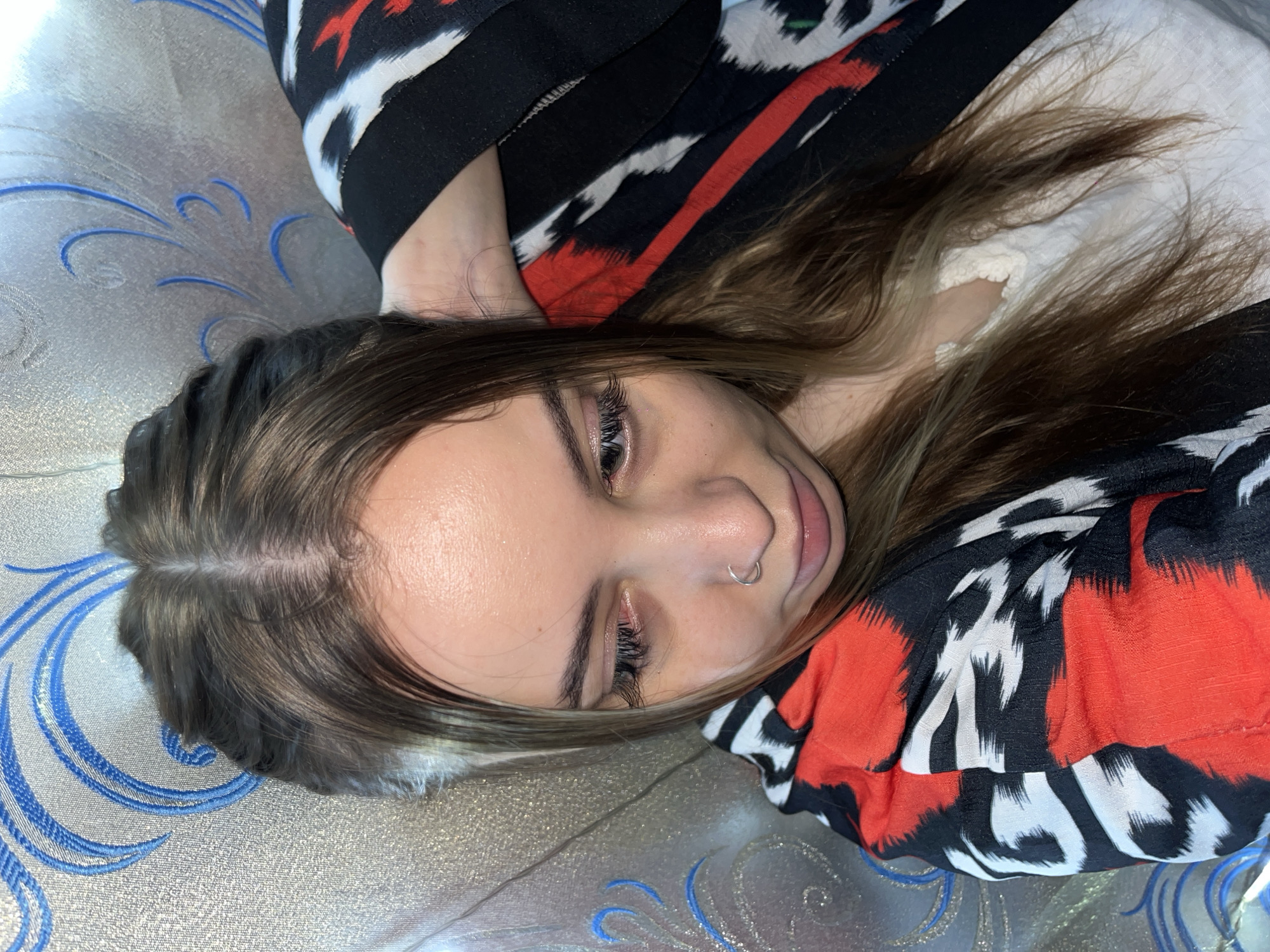 A young woman lies on a patterned surface, possibly a bed or couch, with a red, black, and white patterned garment around her. Her hair is brown with blonde highlights. She has a nose ring. The background is a silver and blue patterned surface. There is no indication of Novopokrovskaya, Russia, or its surroundings.