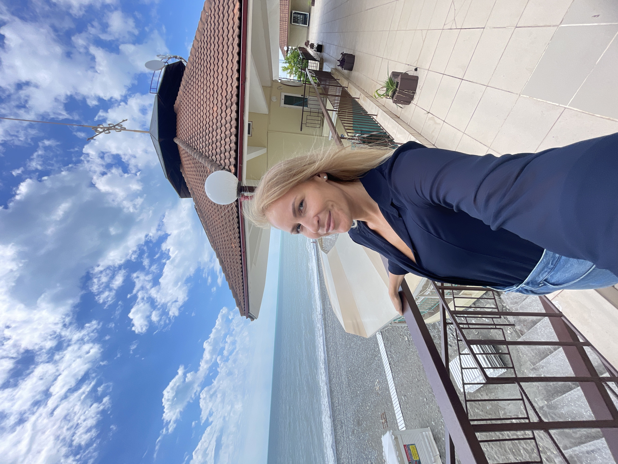 A woman stands on a balcony, overlooking a beach. The woman, fair-skinned with blonde hair, wears a dark blue shirt and jeans. She is positioned on a balcony with brown metal railings and stairs. The balcony is part of a building that is a yellowish color with a brown tile roof. In the background, there is a view of the beach, the sea, and a cloudy sky. The beach appears to be a pebble beach. The image was taken in Sochi, Russia.