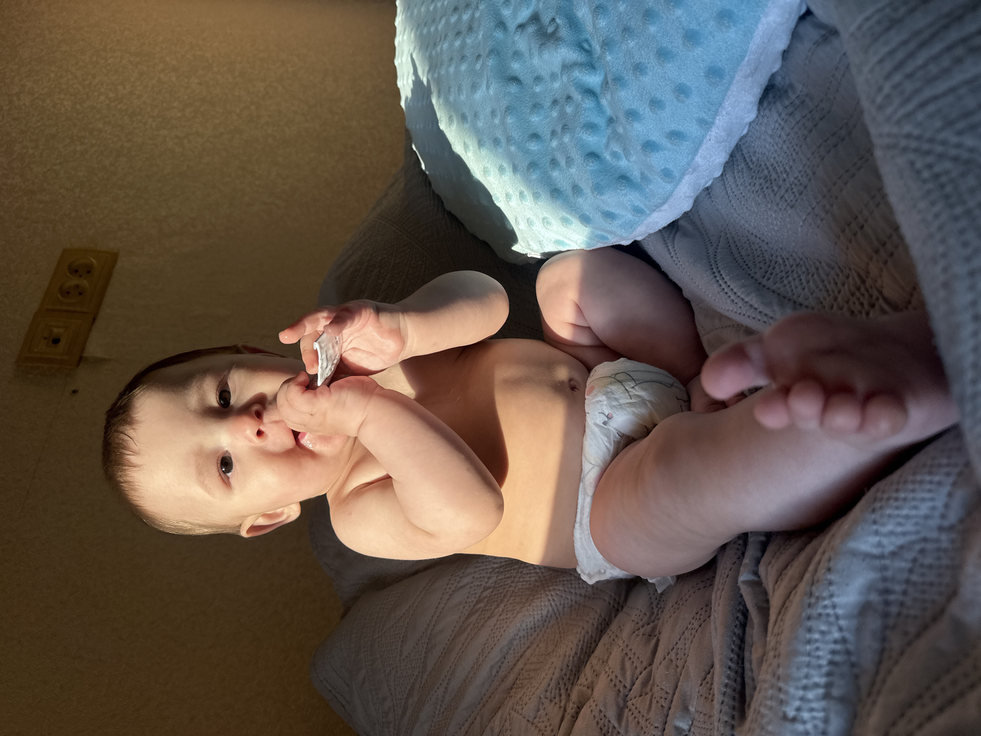 A baby wearing a diaper sits on a grey blanket. The baby is holding a small object in their hands and appears to be chewing on it. The baby has fair skin and dark hair. Sunlight is casting a shadow across the baby's face and body. In the background, there is a textured wall with two electrical outlets. A bright blue, textured pillow is to the right of the baby.